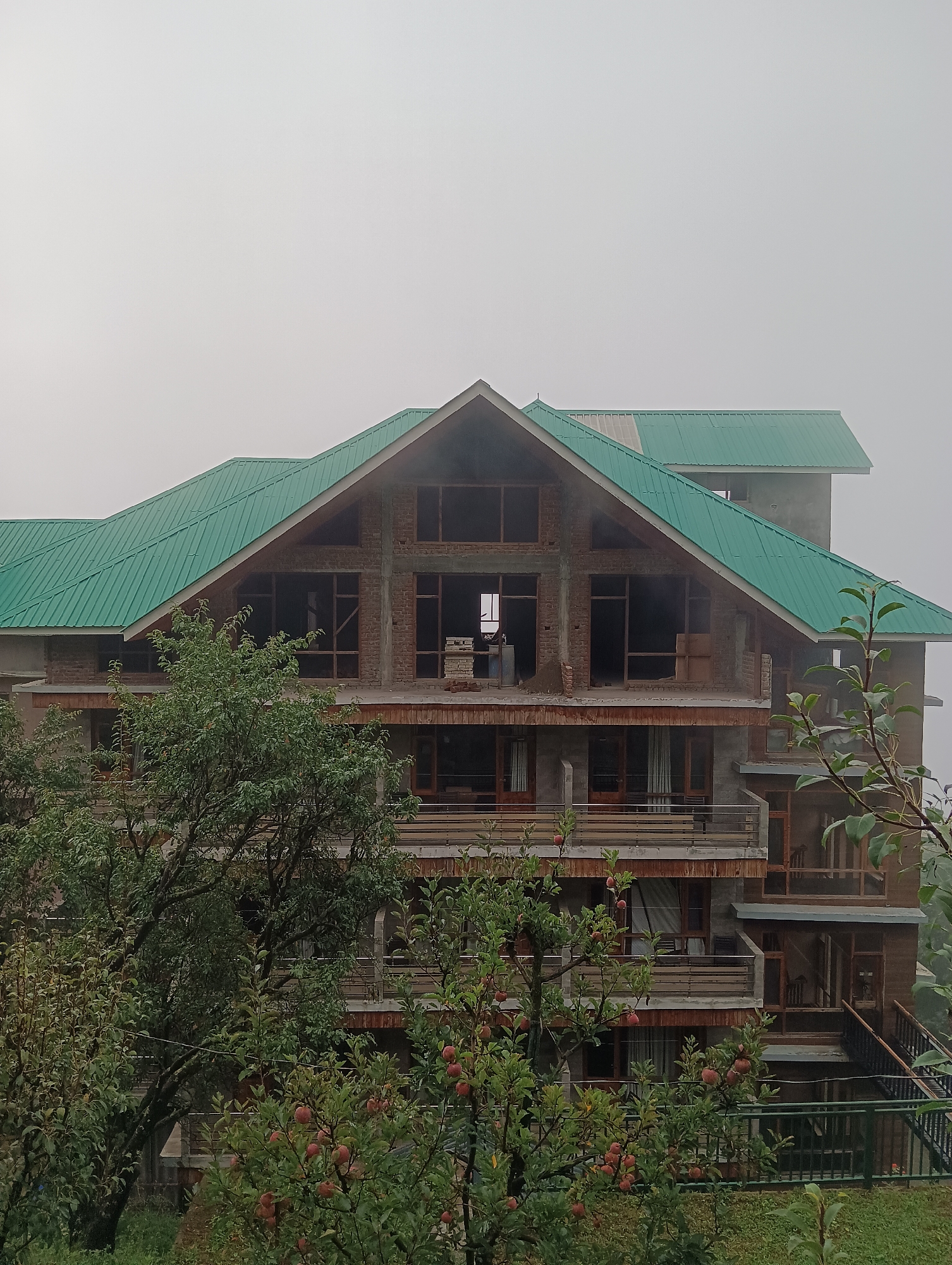 In Chhyal, India, a multi-story building under construction features a prominent, angular green metal roof. The structure is primarily made of brick and concrete, with numerous large, open window frames suggesting the building is not yet complete. Several balconies with railings are visible on different levels. In the foreground, a tree laden with red apples partially obscures the building, with more greenery and foliage surrounding the base. The sky is overcast and hazy, indicating a cloudy or foggy day. No people are visible in the scene.