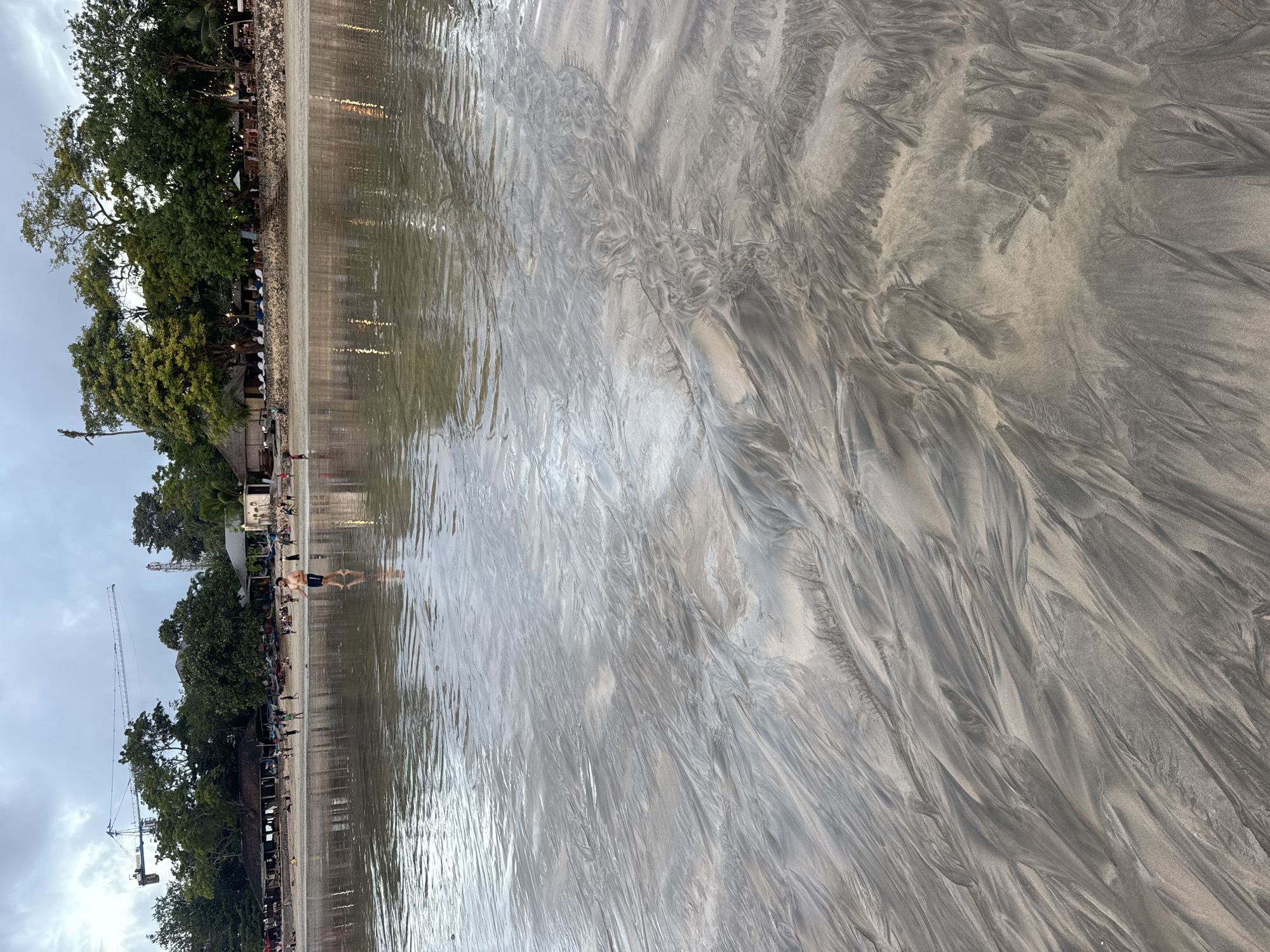 In Kuta Selatan, Indonesia, a serene beach scene unfolds under a cloudy sky. The foreground is dominated by wet sand, intricately patterned by receding tide marks, creating a dynamic interplay of light and shadow. A man, dressed in dark shorts, walks through the shallow water, his reflection visible on the water's surface. In the background, a long stretch of beach is lined with trees and buildings, suggesting a resort or residential area. People are scattered along the shoreline, some standing, others walking, while a few appear to be engaged in activities. A construction crane is visible in the distance, a stark contrast to the natural beauty of the beach. The water is calm, with gentle ripples reflecting the overcast sky and the lights from the buildings along the shore.