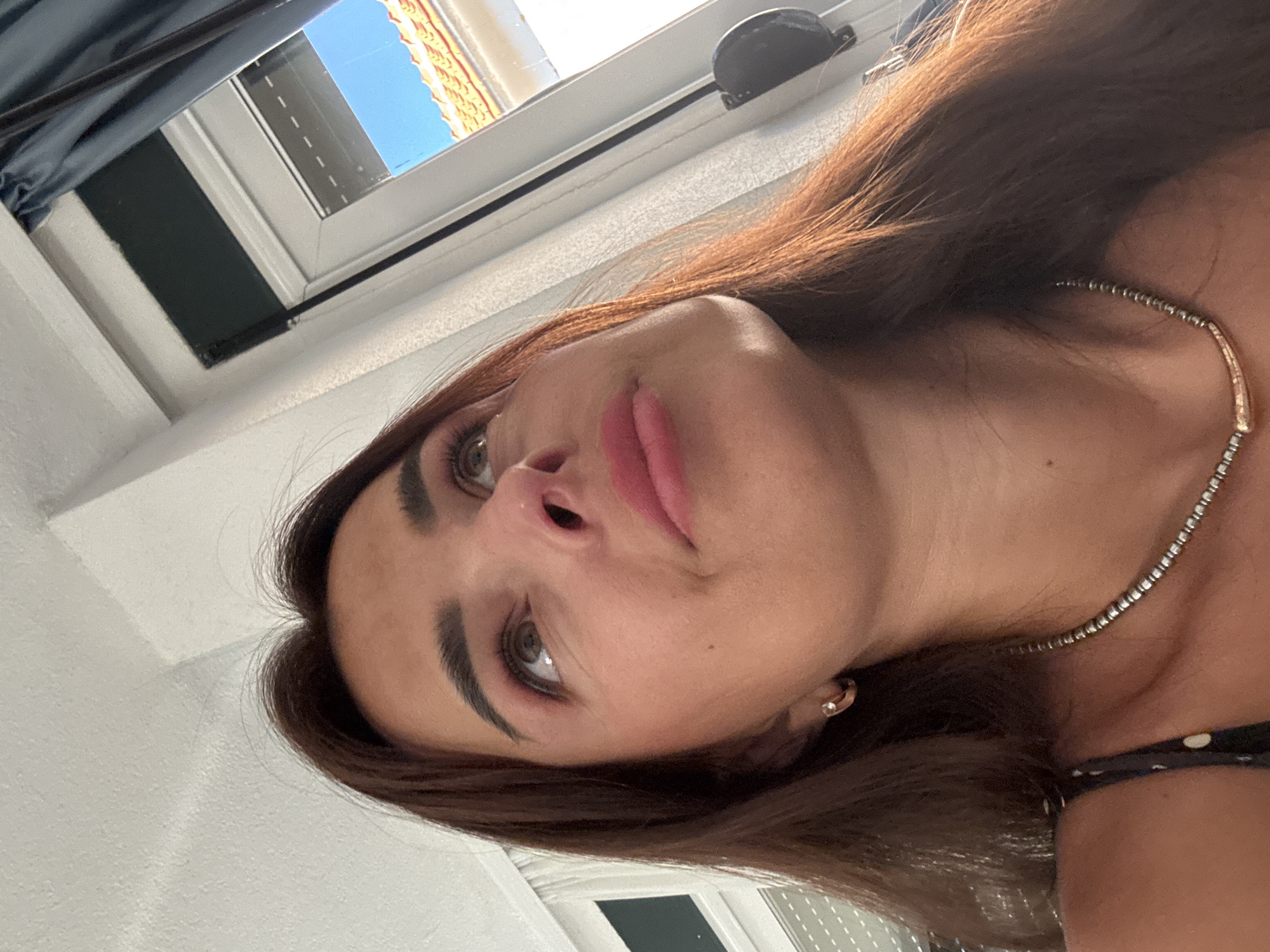 A close-up, low-angle, eye-level shot captures the face of a woman with brown hair, looking upwards towards the right of the frame. She is wearing a dark top with white polka dots and a silver beaded necklace. Her makeup includes pink eyeshadow and lipstick. In the background, a window with blue sky and a tiled roof is visible, along with white curtains and a dark blue curtain. The lighting suggests it is daytime.