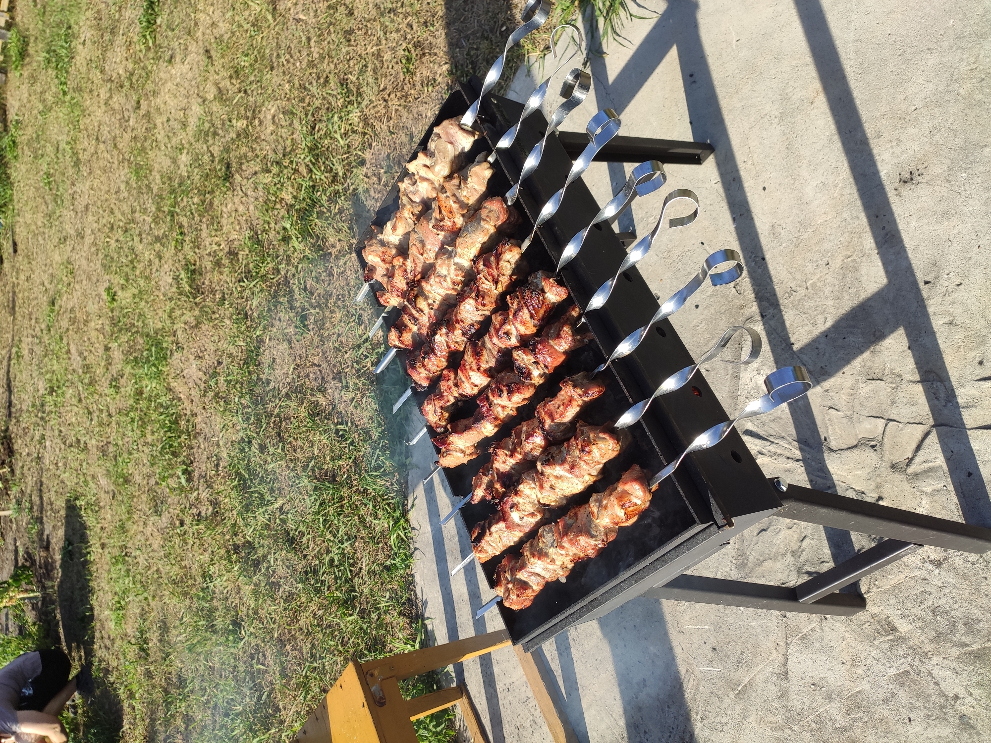 In a residential backyard on a sunny day, a person is crouched near a lawn. In the foreground, a black metal grill is positioned on a concrete patio. Several rows of skewers laden with seasoned meat are cooking over the grill, emitting wisps of smoke. The meat appears to be pork, with some pieces showing charring from the heat. The skewers are made of shiny metal, and some have a distinctive twisted design. The background consists of a grassy area and a wooden table. The image captures a scene of outdoor cooking and relaxation.