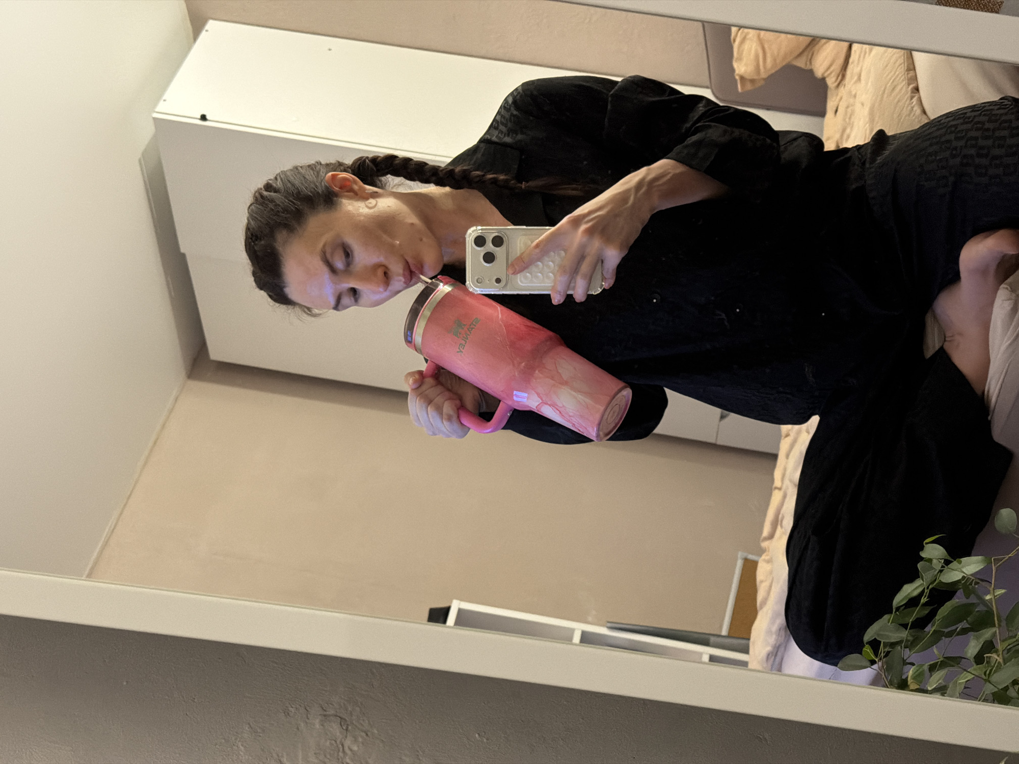 The image depicts a female-presenting individual taking a selfie in a full-length mirror within an interior room, likely a bedroom, located in Perm, Russia. The individual, positioned cross-legged or kneeling, occupies the central foreground of the reflection. She wears dark, possibly black, patterned pajamas consisting of a long-sleeved button-up top and trousers. Her dark hair is styled in braids, one visible over her left shoulder. With her right hand, she holds a large, pink, marbled Stanley tumbler with a handle, bringing a straw to her mouth, appearing to drink. Her left hand holds a silver smartphone with multiple rear cameras, encased in a clear, textured protective cover, directed towards the mirror to capture the image.
The background, visible around and behind the individual's reflection, shows interior furnishings. To the left, a textured dark grey wall contrasts with a smoother light beige or pale pink wall. A white, two-door wardrobe or cabinet with dark fasteners is visible behind her head and right shoulder. To the right, a bed with a dark grey headboard, a beige duvet, and a pillow is partially visible. A white shelving unit is reflected on the left side of the mirror. In the lower left foreground, a green leafy houseplant is partially visible, indicating its placement near the mirror. The ceiling is white.