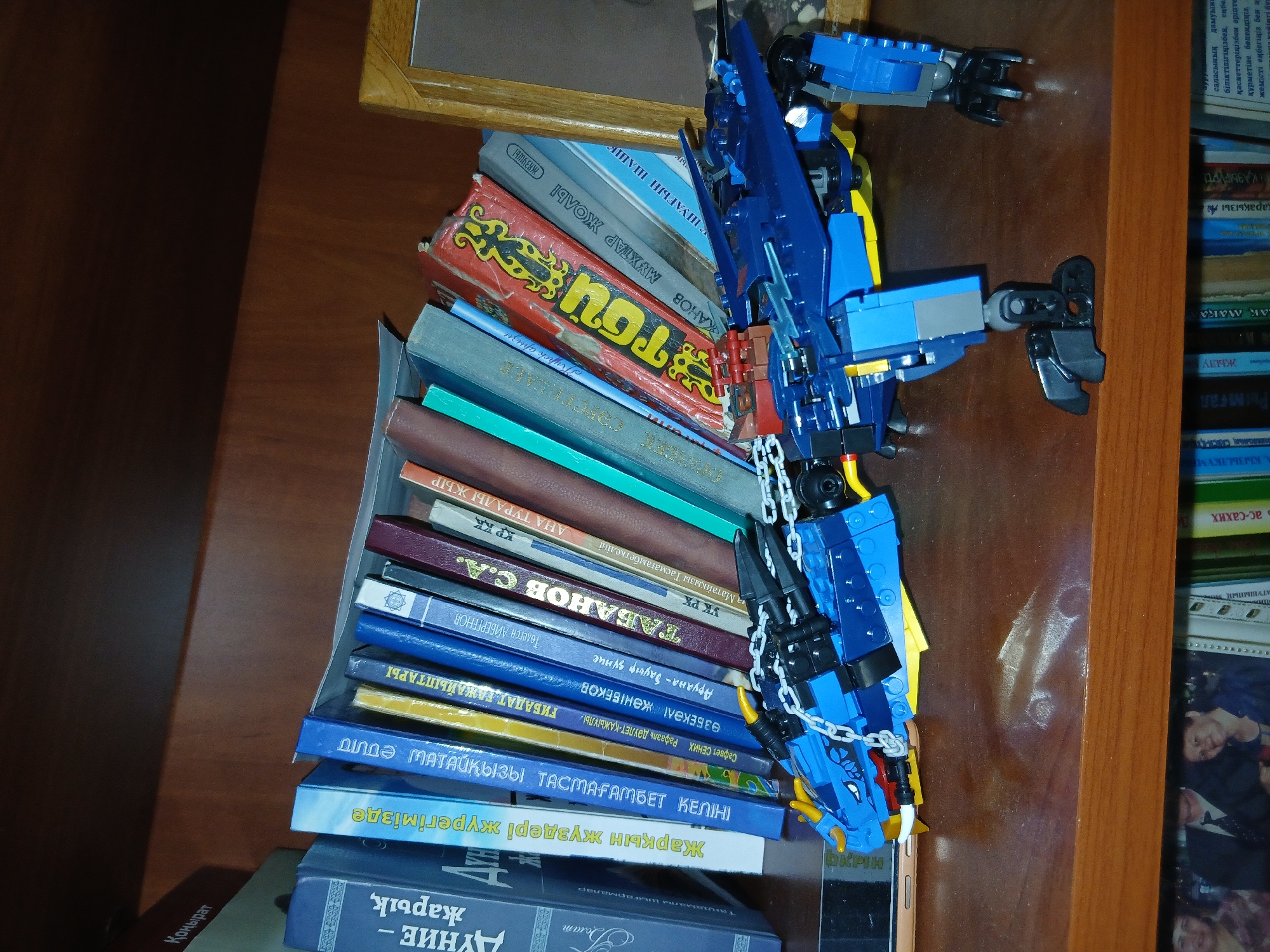 The image shows a bookshelf filled with books and a blue Lego dragon model. The books are arranged at an angle, leaning against each other. The Lego dragon is placed on the shelf in front of the books, with its head facing left. The environment appears to be indoors, likely a home or library. There are no people visible in the image. The Lego dragon has chains attached to its head and neck. The bookshelf is made of wood. The background of the image is out of focus, but it appears to be a wooden wall. The lighting is dim, suggesting it may be evening or the room is not well-lit. The books have various titles written in Cyrillic script. The overall scene is a still life composition with a toy and books as the main subjects.
