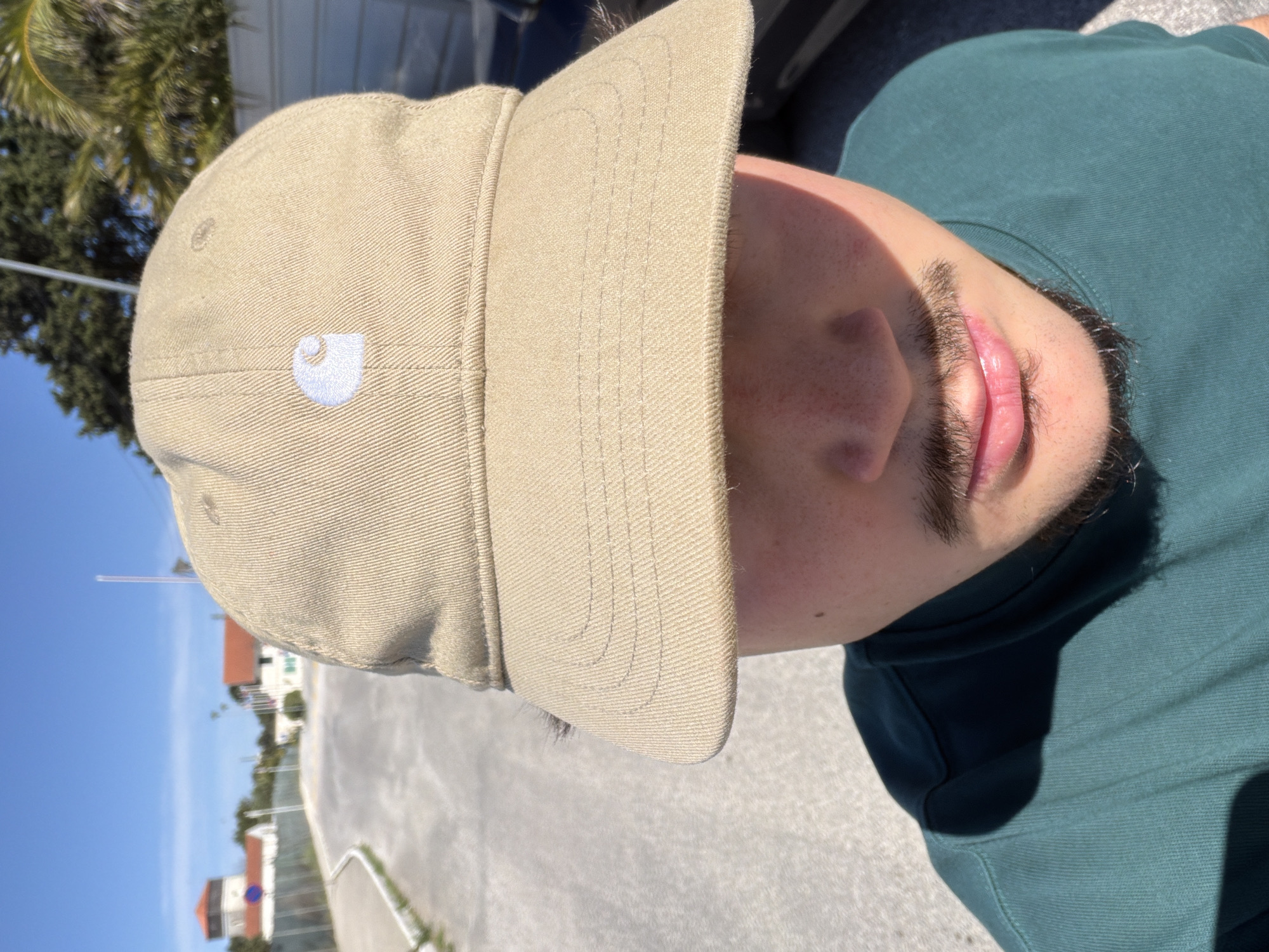A close-up photograph depicts a person wearing a tan Carhartt cap and a dark green t-shirt. The brim of the cap obscures much of the person's face, revealing the nose, mouth, and beard. The background shows a street with buildings, a white fence, and a clear blue sky, indicating an outdoor setting. The location is likely in Cascais, Portugal, based on the architectural style of the buildings and the landscaping.
