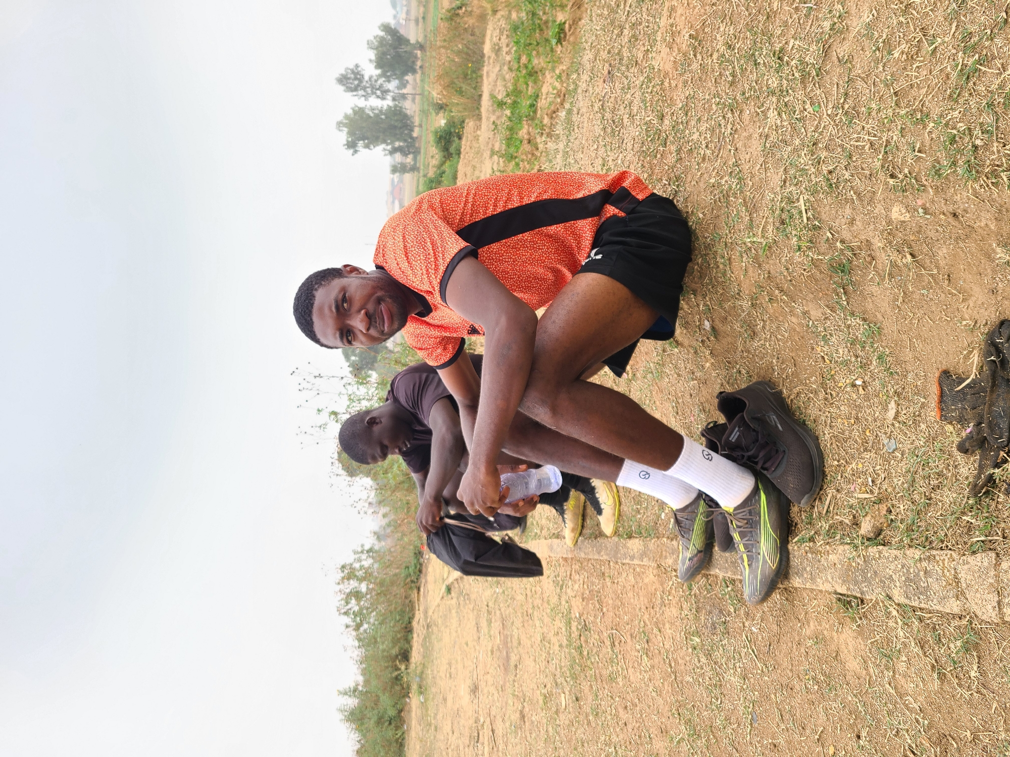 Two young men are outdoors in Barikin Jirga Summa, Nigeria, likely in a field. One man in the foreground is wearing an orange patterned shirt with black accents and black shorts. He is seated with his knees up and smiling at the camera. He is wearing white socks and running shoes. Behind him, another man in a dark shirt is seated, looking down. A water bottle and a bag are near him. Running shoes are seen near both men. The ground is dry with sparse grass. The sky is overcast.