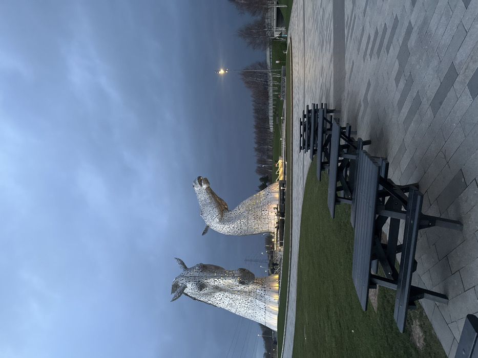 The image displays an outdoor scene at The Helix park in Falkirk, United Kingdom, at twilight or under heavily overcast conditions. The primary subjects are two monumental, highly detailed metallic horse head sculptures, known as The Kelpies, positioned centrally in the background and illuminated from below by warm-toned lights. The left sculpture depicts a horse's head facing forward, while the right sculpture shows a horse's head raised with an open mouth. Both are constructed from numerous faceted metal plates. In the foreground and midground, a linear arrangement of approximately six dark-colored wooden picnic tables with attached benches occupies a gray patterned paved walkway. To the left of the walkway is a maintained green grass area. The background features a line of bare deciduous trees, a street lamp emitting light, power lines, and elements of distant infrastructure including a bridge and low walls. The sky is dark and predominantly covered by dense, grey clouds. No individuals, actions, or interactions are present in the scene.