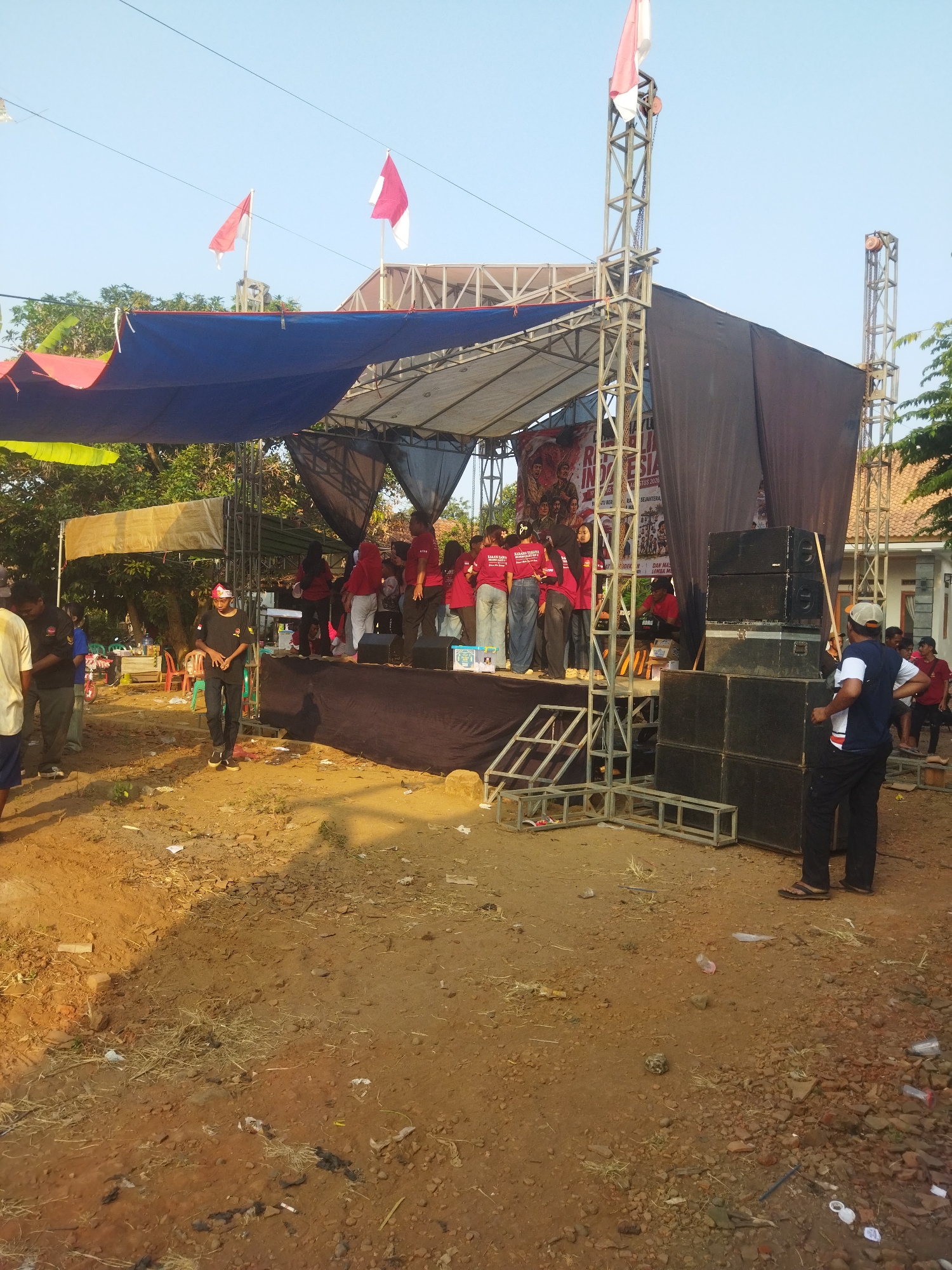 The image depicts an outdoor stage set up on a dirt lot, likely for a community event in the city of Banjar, Indonesia. The stage has a blue and red tarp overhead supported by metal scaffolding. Two Indonesian flags, red and white, are visible flying above the stage. A banner with text in Indonesian is hung behind the performers on stage.
On the stage, several people, mostly wearing red shirts, are gathered. Some appear to be performers or organizers. In the foreground, on the right side of the image, a stack of large black speakers is arranged. A man wearing a blue shirt and a cap stands near the speakers, facing the stage. To the left of the stage, another man in a black shirt and a red headband is walking. The ground is covered with dirt, scattered debris, and some dry grass. In the background, trees and a building with a tiled roof are visible. The sky is clear and blue. The overall scene suggests a festive or celebratory gathering.