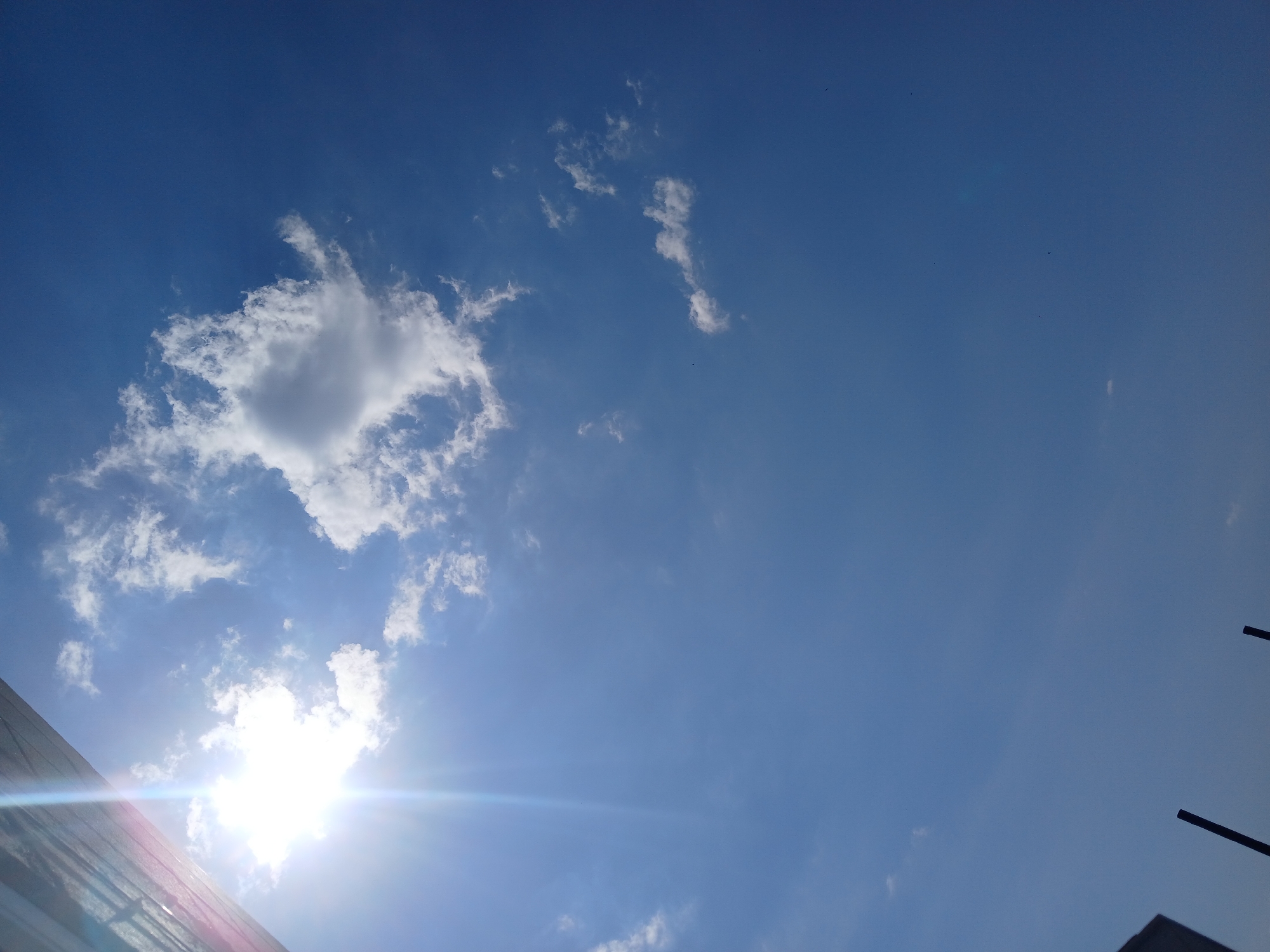 The image shows a bright blue sky with scattered white clouds. The sun is visible in the upper left corner, creating a lens flare. In the lower left and bottom center of the image, there are dark, vertical objects that appear to be poles or structures. The image was taken in the city of Trușeni, Moldova.