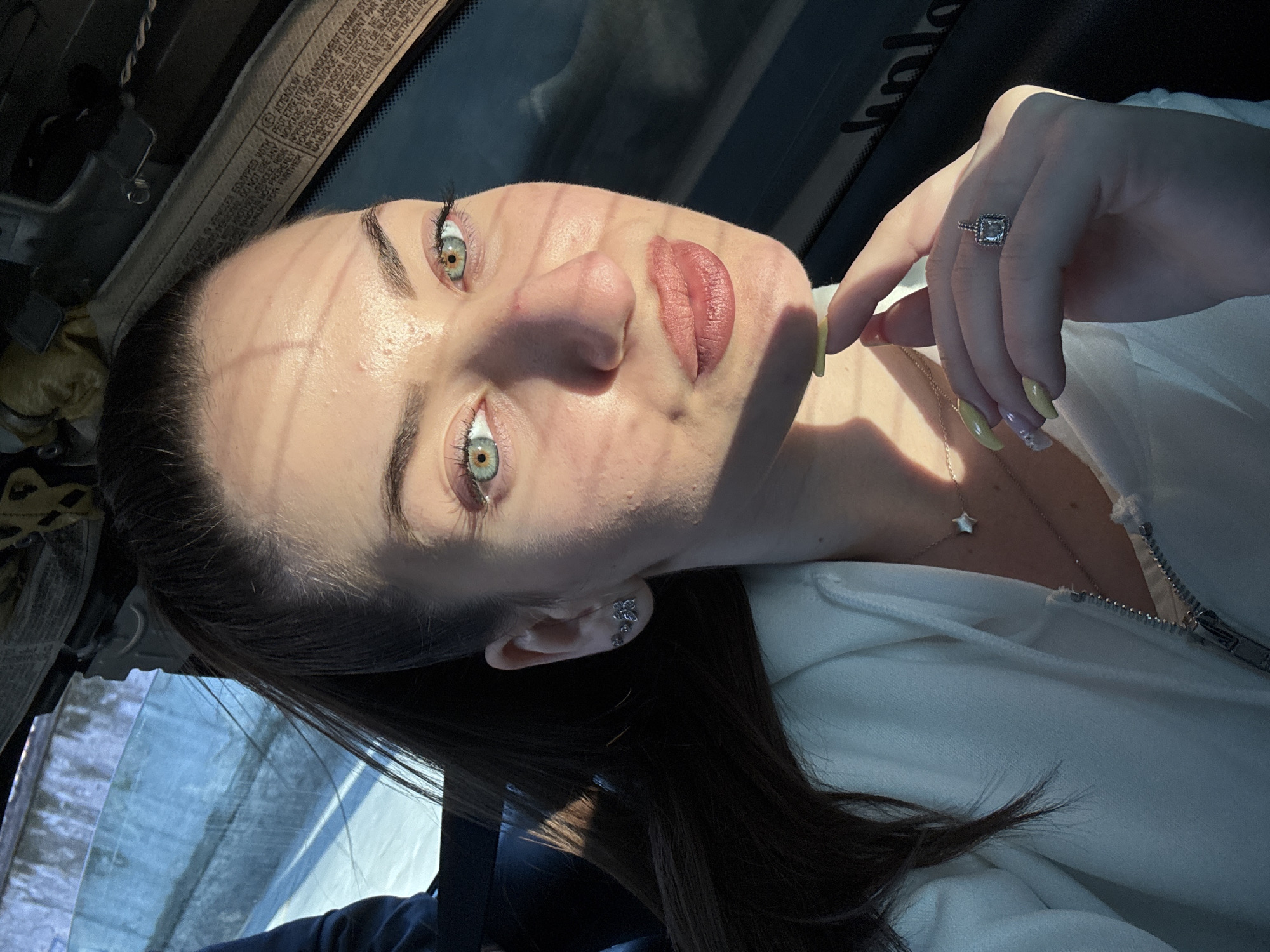 A selfie taken inside a vehicle. The image shows a woman with light skin, dark hair, and hazel eyes. She is wearing a white jacket and a necklace with a star-shaped pendant. She is wearing earrings in her left ear and a ring on her left hand. The woman is looking directly at the camera with a slight smile. The lighting suggests she is sitting in the car during the day. The interior of the car is visible, including parts of the dashboard and the window.
The location in the city of Plovdiv, Bulgaria is not determinable from the image.