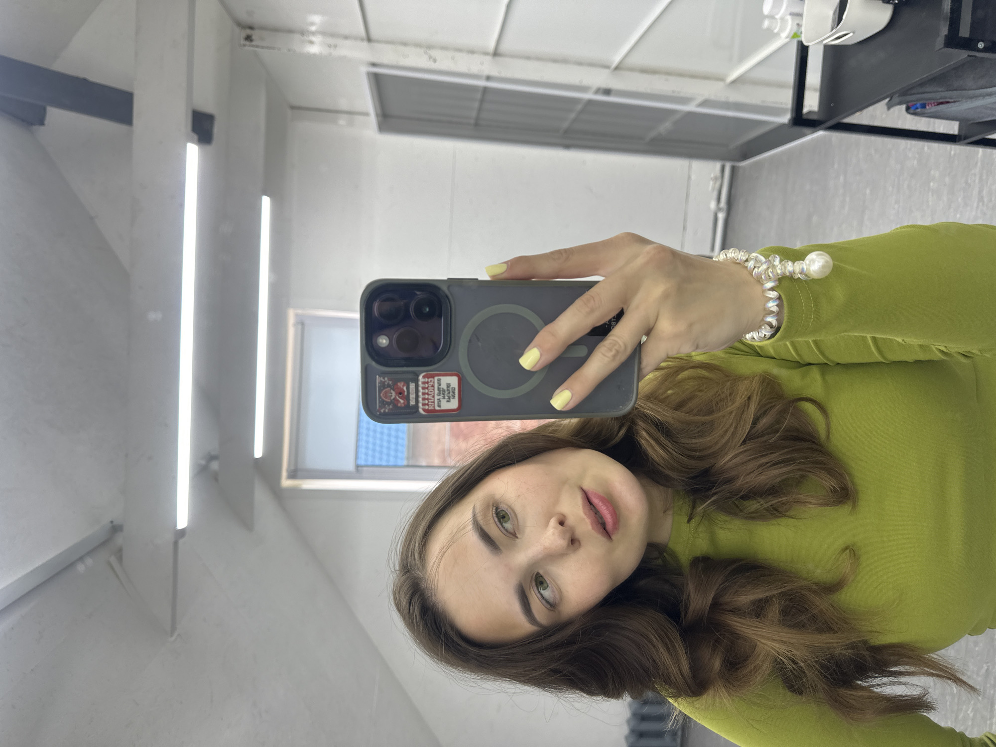 A young woman is taking a selfie in a room with bright overhead lighting and white walls. She has long, wavy brown hair and is wearing a lime green long-sleeved shirt. On her right hand, she is wearing a pearl bracelet. She is holding a dark-colored smartphone, which is reflecting some of the room's interior. The room appears to be a salon or a similar commercial space, as indicated by a shelf unit with bottles on the right side of the image. The overall impression is casual and contemporary.