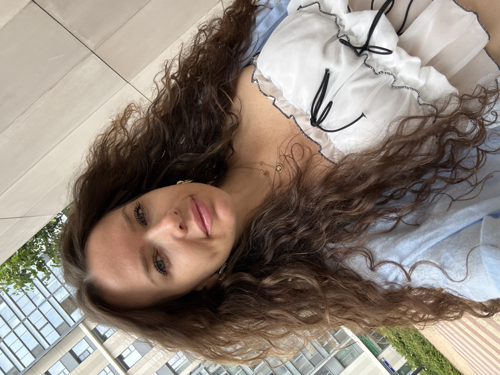 A woman with long, curly brown hair is captured in a selfie outdoors in front of a modern building in Khimki, Russia. She is wearing a white off-the-shoulder top with ruffled trim and black ties, layered over a light blue garment. Her makeup is subtle, with a focus on her eyes and lips. The background features a section of a glass-fronted building with a grid-like structure, suggesting an urban setting. Adjacent to the building, there is a glimpse of greenery and a striped walkway. The overall scene suggests a casual, daytime setting in a city.