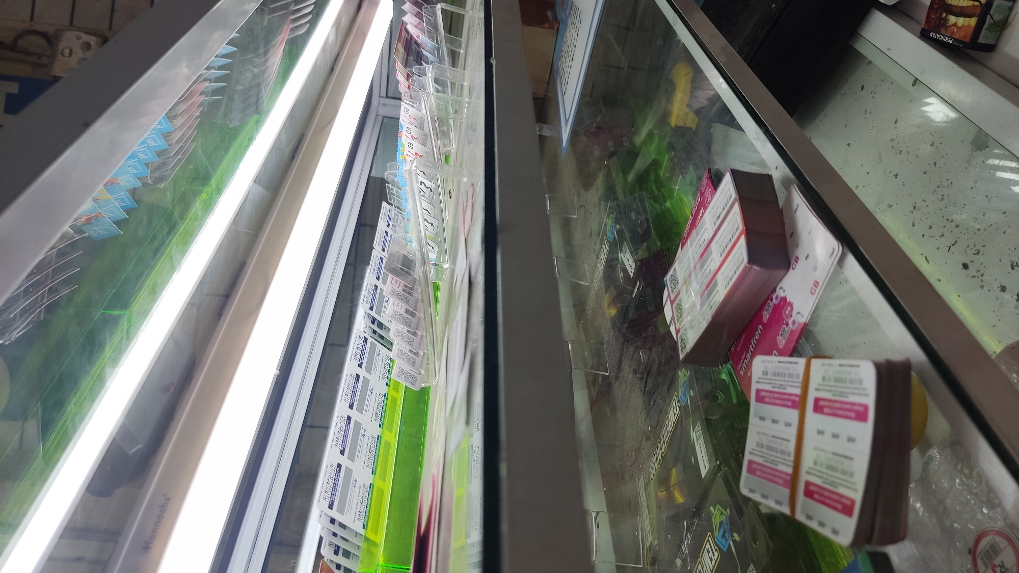 The image provides an angled, top-down view into a brightly lit retail display case, characteristic of a shop or kiosk in Lhokseumawe, Indonesia. The display case is made of glass and features prominent fluorescent lighting.
On the left side of the case, numerous rectangular cards, likely SIM cards or mobile top-up vouchers, are neatly arranged in clear plastic dispensers. Visible text on these cards includes brand names such as "IM3 Ooredoo" and "Smartfren," along with numerical values (e.g., "Rp") and "GB" (indicating Gigabytes for data plans). Many cards also display QR codes and barcodes. Bright, reflecting fluorescent tubes, one bearing the brand "Hannochs," illuminate this section. A green surface is visible beneath the cards.
The right side of the display case contains more mobile phone-related products. A stack of reddish-brown boxes, also branded "Smartfren" with QR codes and text, is prominently placed. Below these, a bundle of pink and white cards, secured with a rubber band and marked "GB," rests on the glass surface. Other items visible include a white cylindrical object, some clear bubble wrap, and a partially obscured brown package with the Indonesian word "PERINGATAN" (WARNING).
The scene depicts an organized retail setting, likely a telecommunications store or a general store selling mobile phone accessories and services. No individuals are present in the image. The artificial lighting suggests an indoor environment, making the time of day indiscernible.