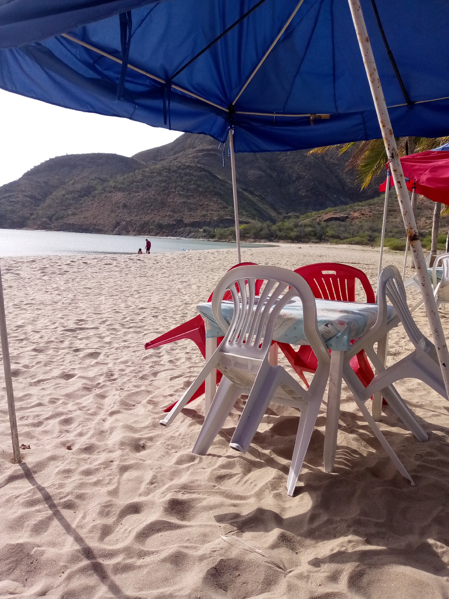 The scene is set on a sandy beach in Juan Griego, Venezuela. A blue beach umbrella dominates the upper portion of the frame, providing shade to a plastic table covered with a patterned light blue tablecloth. Red and white plastic chairs surround the table. In the background, a bay is visible with mountains in the distance. Two individuals are standing near the water's edge. The lighting suggests it is daytime with strong sunlight casting shadows on the sand. Another red umbrella is visible on the right. The sand is textured with footprints and shadows.