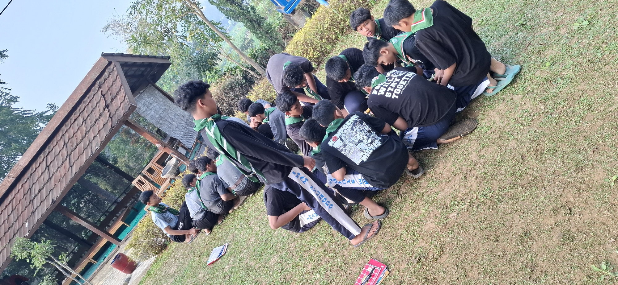 In the city of Puntukrejo, Indonesia, a group of young males, presumably scouts based on their green neckerchiefs, are gathered outdoors on a grassy area. They are sitting and kneeling in a circle, with some leaning in towards the center. Many are wearing dark t-shirts, and at least one individual is wearing athletic pants with white and blue stripes. The ground is covered in a mixture of grass and dirt. In the background, there is a wooden structure with a tiled roof, possibly a pavilion or shelter. Lush green trees and foliage surround the area, suggesting a natural or park-like setting. The sky is visible and appears to be clear and bright. One scout in the foreground has text on his t-shirt that appears to read "FOR WILL STAY TOGETHER".