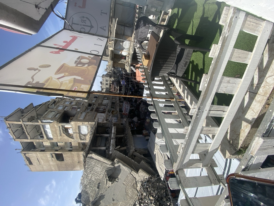 The image captures an elevated view of a severely damaged urban environment in Khan Yunis, Palestinian Territory, under clear daylight conditions. In the immediate foreground, a white wooden picket fence defines an outdoor seating area furnished with artificial green turf, a dark woven chair, and a small table supporting a wooden box. A portion of a smartphone is visible in the bottom left corner. Above, two commercial signs are mounted on poles; one large sign displays "Titanic" with an anchor logo and Arabic script, while another features graphics of a coffee grinder, a traditional Arabic coffee pot (dallah), and a mug.
Beyond this foreground structure, a busy street or market area is visible, crowded with pedestrians and numerous stalls covered by light-colored awnings, displaying goods. This thoroughfare is flanked by extensively damaged buildings. To the left, a large multi-story concrete building is partially collapsed, with significant structural failure evident in its upper sections and a substantial amount of rubble at its base. In the background, a tall, multi-story concrete building stands prominently, exhibiting severe structural damage, including exposed internal framework, missing exterior walls, and debris within window openings. Further into the distance, additional damaged urban structures line the horizon beneath a clear blue sky.