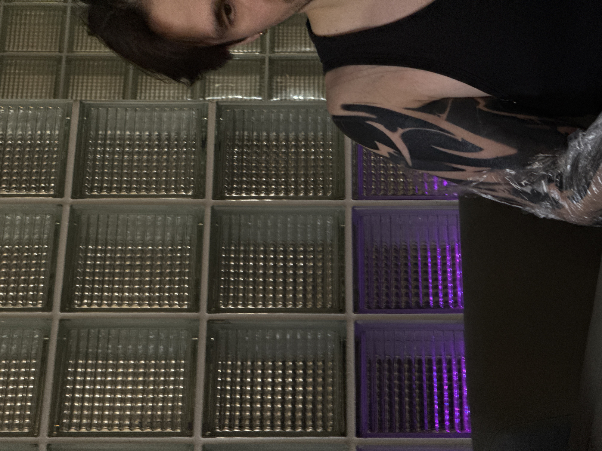 The image captures a partial, downward-angled view of a person, likely male, with dark hair, looking towards the camera. He wears a black top, and a distinctive black, tribal-style tattoo is visible on his right arm. This tattoo appears to be freshly done, as it is covered in clear plastic wrap.
The background consists of a wall constructed from textured glass blocks. Most of these blocks are clear or frosted, reflecting ambient light. However, several blocks on the right side of the wall are illuminated with a vibrant purple light, creating a striking contrast. The textured surface of the glass blocks is prominent, with light glinting off their ridges.
The setting is indoors, with artificial lighting accentuating the glass block wall. The person's expression is neutral as they look directly at the viewer. There is no visible text in the image. The scene could be an interior space in Moscow, Russia, characterized by its modern design elements and unique lighting.