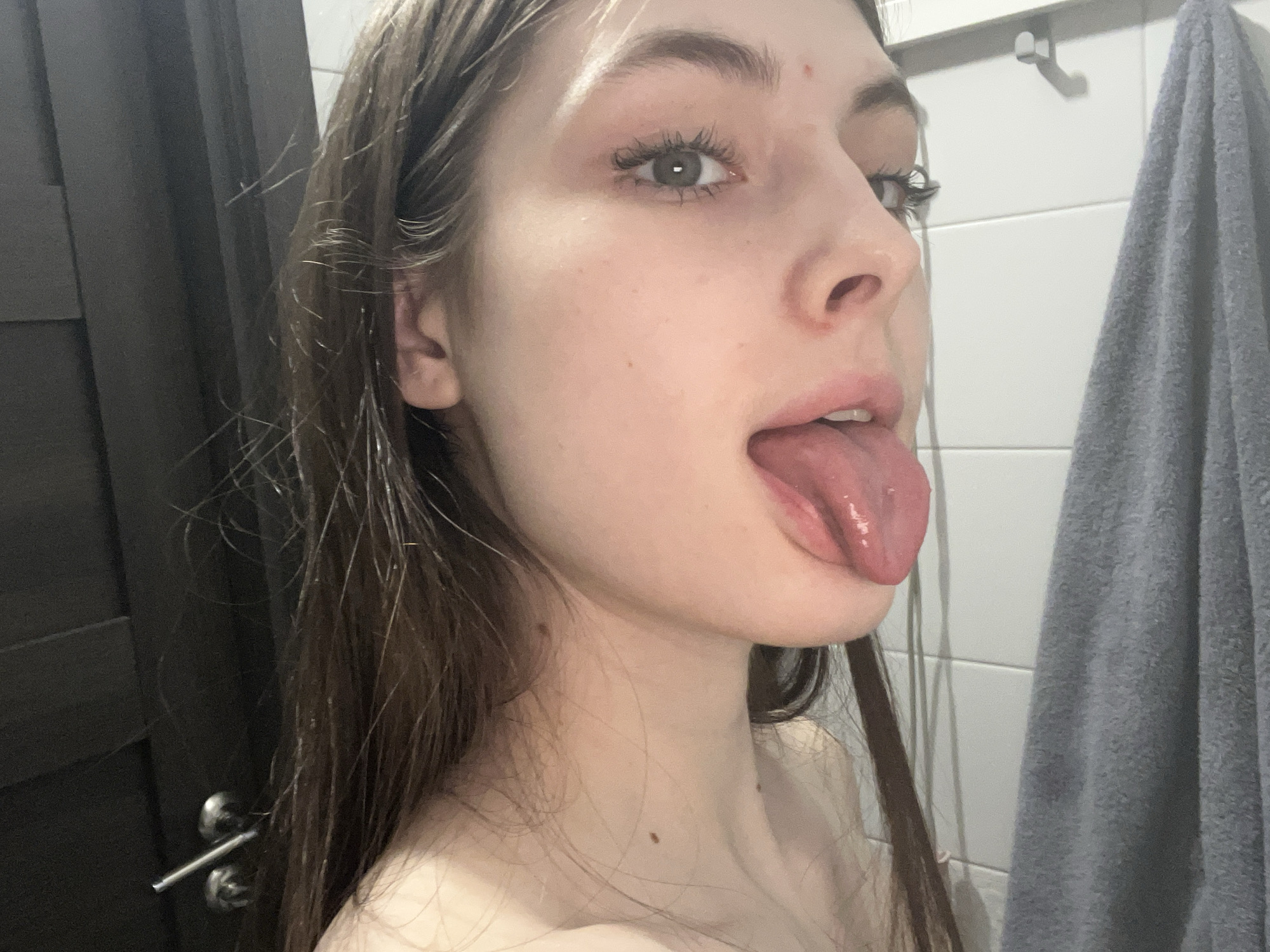 A young woman is positioned in a bathroom. She has light skin, gray eyes, and long brown hair. Her mouth is open and her tongue is extended. To her left, there is a dark-colored door with a silver handle. Behind her, there is a white tiled wall with a silver towel hook and a gray towel. No indications suggest this scene is in Saint Petersburg.