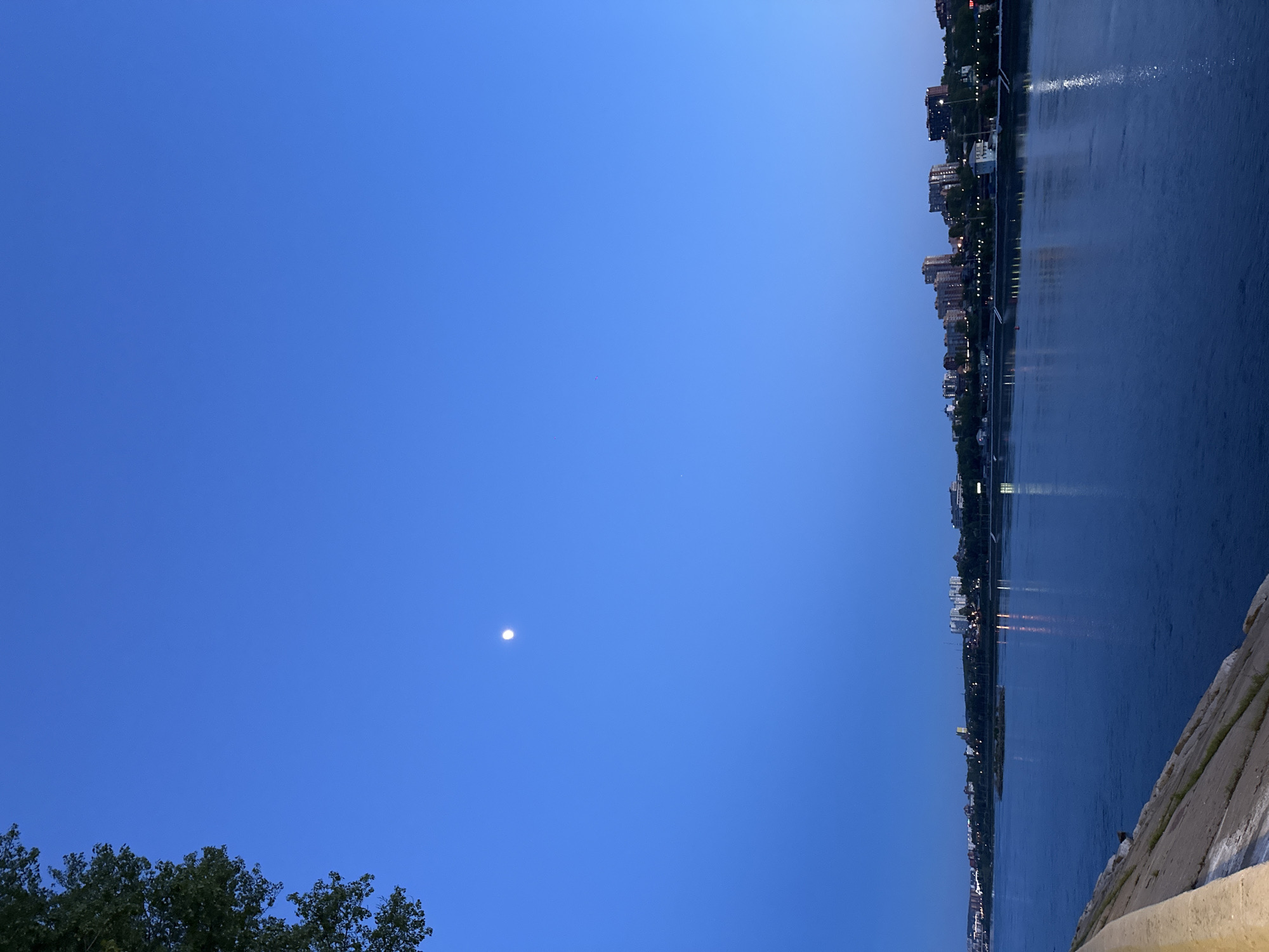 The image shows a nighttime view of the Angara River in Irkutsk, Russia. The sky is a deep blue with a small, bright moon visible. The river reflects the city lights and sky. The city skyline, with buildings of varying heights, stretches along the riverbank. In the foreground, there is a partial view of some trees and concrete structures.