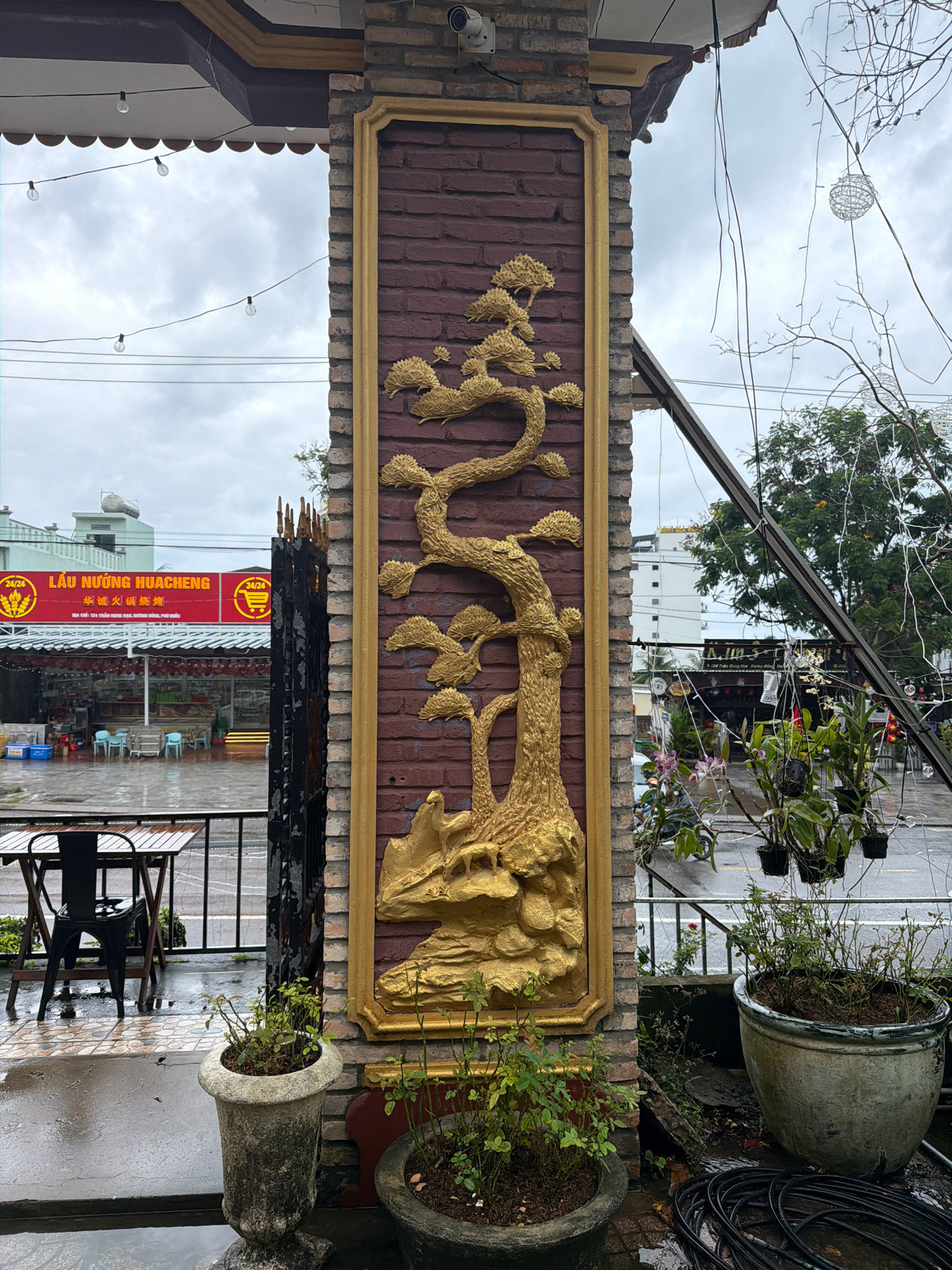 The image depicts an outdoor scene in Phú Quốc, Vietnam, likely a restaurant entrance. A brick pillar adorned with a golden relief of a bonsai tree stands prominently. To the left, a black metal gate opens to a courtyard with a black metal table and chairs set on a tiled, wet surface, indicating recent rain. A red awning with Vietnamese text is visible in the background, advertising "LẨU NƯỚNG HUACHENG." Beyond the awning, a building with a rooftop water tank and a sign reading "A.LIM'S" is visible. On the right side of the pillar, several potted plants hang, and a large pot sits on the ground, with black electrical cords coiled nearby. The sky is overcast, and the overall atmosphere suggests a slightly gloomy, damp day.