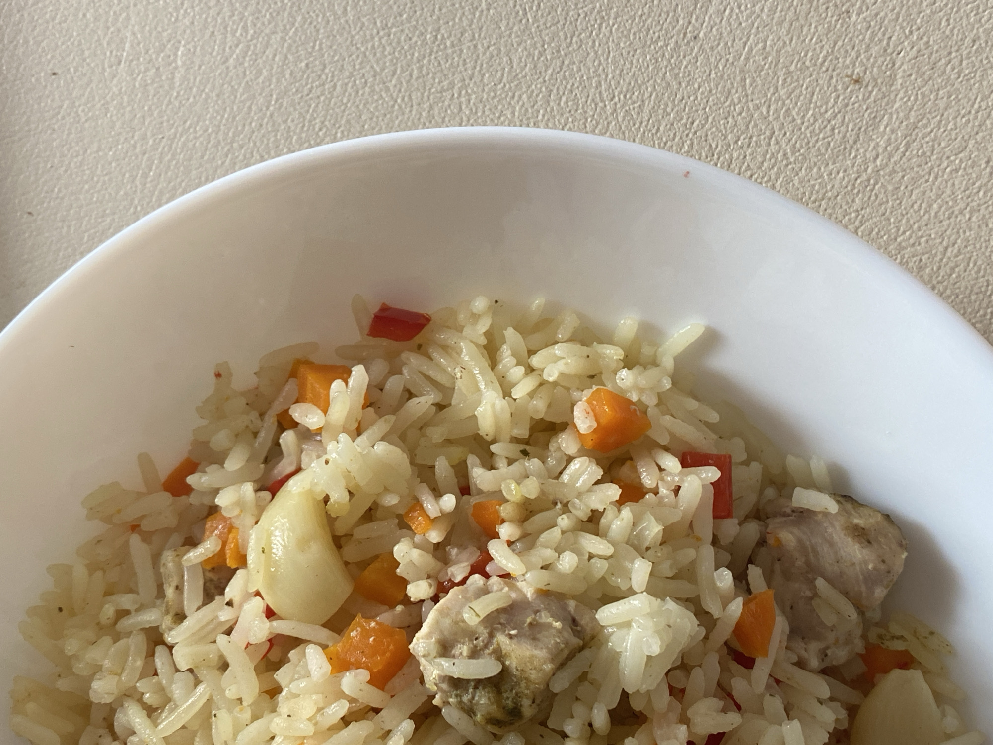 A close-up, overhead shot shows a white bowl filled with pilaf. The pilaf contains rice, chunks of meat, diced carrots, and what appears to be garlic cloves. The bowl is placed on a light-colored, textured surface. The lighting suggests it is daytime, with natural light coming from the right. There is no indication of the location being in Saint Petersburg, Russia.