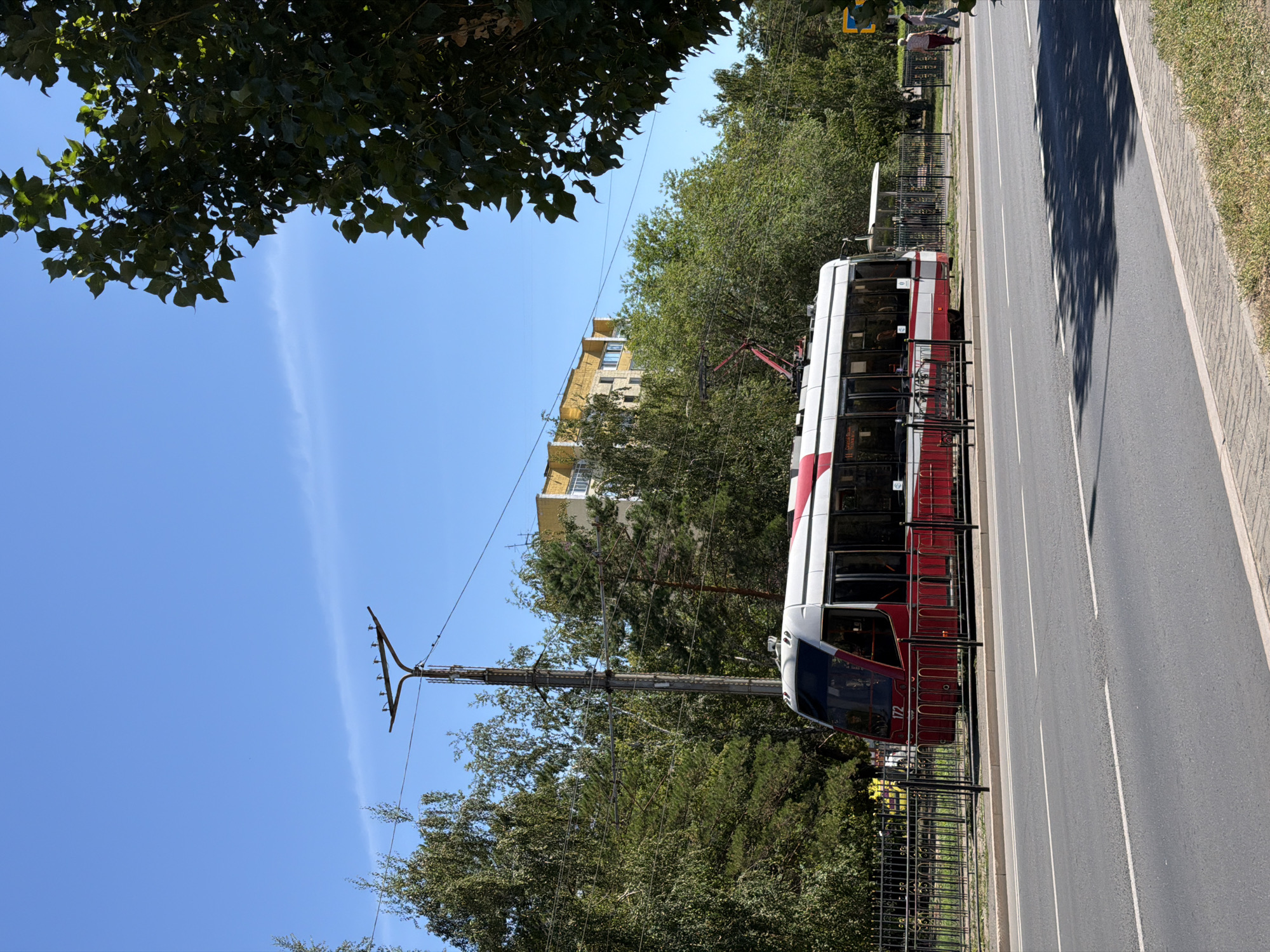A modern red and white tram, numbered 172, is stopped at a tram stop. The tram has large windows and a sleek design. It is positioned on a tram track that runs alongside a road. In the background, there are several trees, a multi-story residential building with yellow facades, and a utility pole with wires. The sky is clear blue. Two people are walking away from the tram stop on the sidewalk. The scene appears to be a typical urban streetscape in Pavlodar, Kazakhstan, during daylight hours.