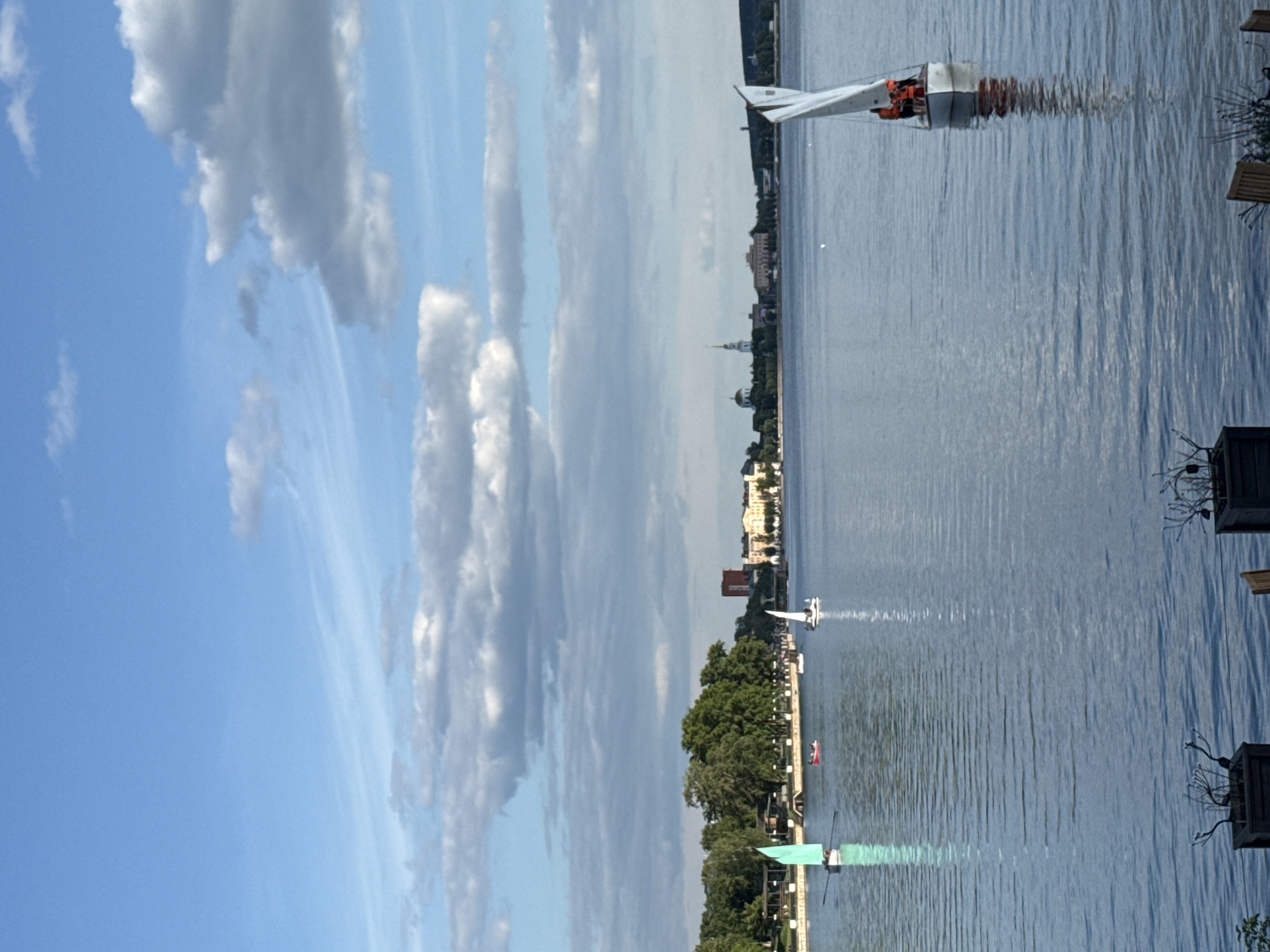 This image presents a detailed view of the Votkinsk Pond in Votkinsk, Russia, under a partly cloudy sky. The upper approximately one-third of the frame displays a light blue sky with numerous scattered white and light grey clouds, ranging from thin, wispy cirrus-like formations to more defined cumulus clouds, with sunlight illuminating the scene.
The central and lower portions of the image are dominated by the calm, reflective surface of the Votkinsk Pond, which appears a deep blue-grey due to reflections of the sky and surroundings, exhibiting subtle ripples.
In the immediate foreground, at the very bottom edge, two dark, rectangular planters with decorative twig-like elements are partially visible, along with a small portion of a wooden chair, suggesting a vantage point from a lakeside cafe or park.
Multiple boats are present on the water. In the mid-ground, on the left side, a light green sailboat with a single white sail is being propelled by visible oars, with at least one person operating it. Further to the right and slightly behind, a small red boat, possibly a paddleboat or rowboat, contains two occupants. Another small white sailboat with a single sail is visible further right in the mid-ground.
Prominently positioned in the lower right foreground, a white sailboat with a triangular white sail is actively underway. Three individuals are visible within this sailboat, each distinctly wearing a bright orange life vest, indicating active sailing. The boat casts a clear, elongated reflection on the water's surface.
The distant shoreline across the pond, spanning from left to right, features a dense line of green trees. Beyond these trees, the cityscape of Votkinsk is visible. Notable structures include a prominent reddish-brown building on the far left, followed by a series of lighter-colored buildings. Towards the right-center of the distant skyline, two distinct structures with spires or domes, characteristic of churches or historical buildings, stand out against the horizon. The land beyond the city appears to gently rise.