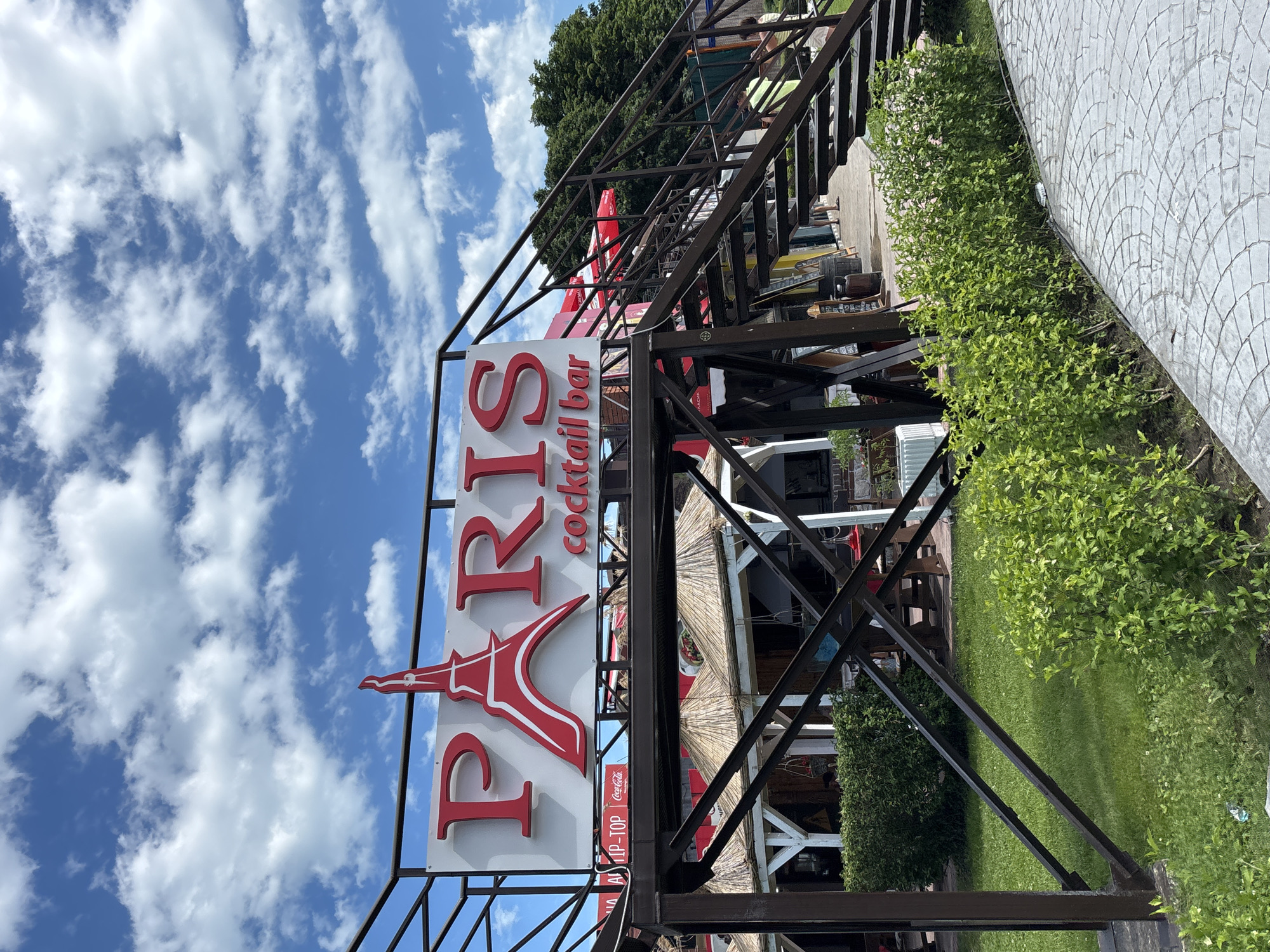 This image, taken in Varna, Bulgaria, shows a sign for a cocktail bar named "PARIS." The sign features a large red "PARIS" with a silhouette of the Eiffel Tower integrated into the letter "A." Below "PARIS" in smaller red text, it reads "cocktail bar."
The sign is mounted on a dark metal frame, which also supports an outdoor seating area or structure. In the background, there is lush green foliage, a lawn, and a paved area with a decorative pattern. The sky is blue with scattered white clouds, suggesting a bright and possibly sunny day.
Although people are present in the background, they are not the main focus and are not clearly identifiable. There are some tables and chairs visible, along with what appears to be a thatched roof structure, indicating an outdoor bar or restaurant setting. A Coca-Cola logo is also visible on a smaller sign. The overall scene appears to be a relaxed outdoor environment, possibly during daylight hours.