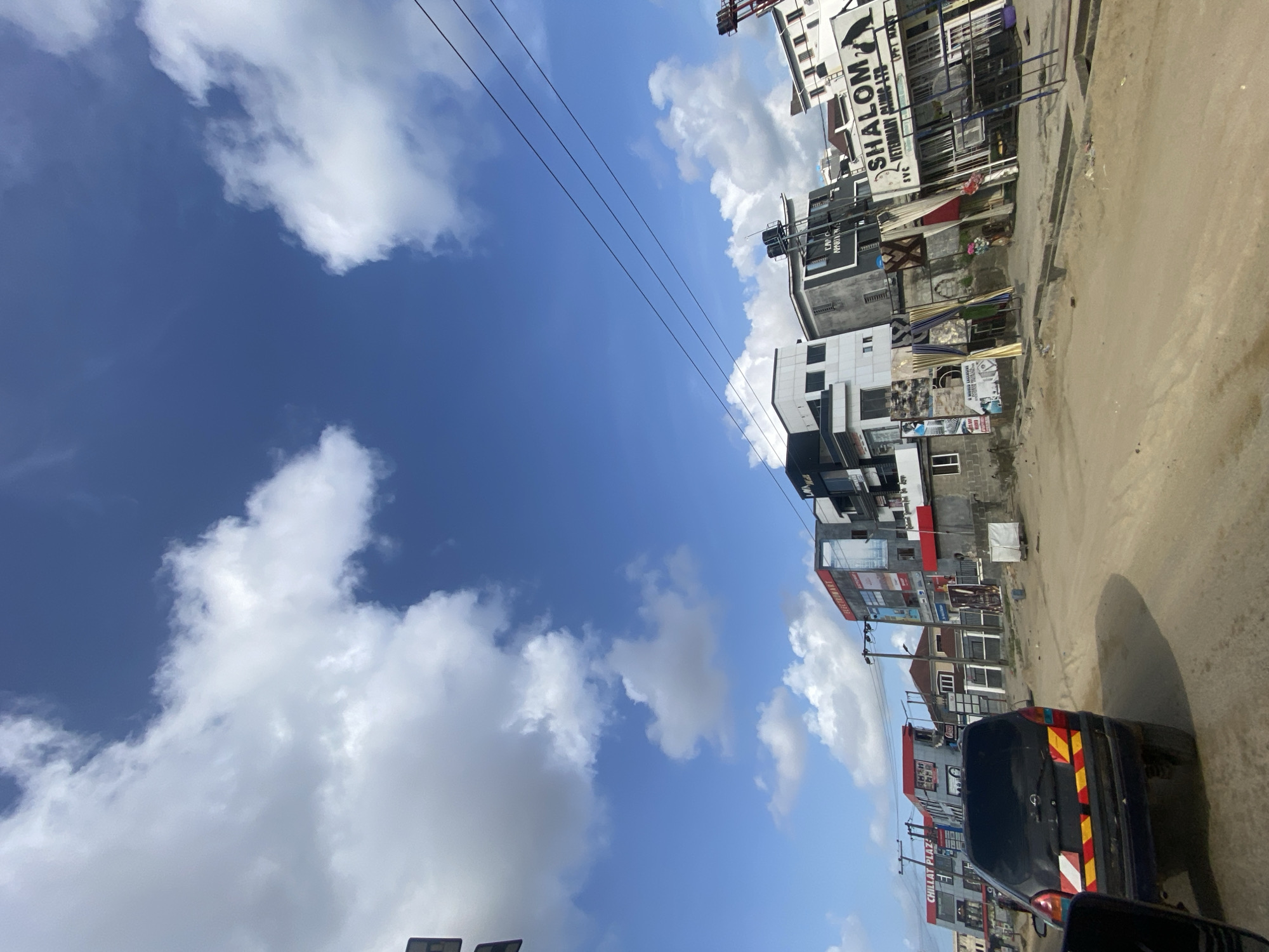 The image shows a street scene in Lagos, Nigeria. The view is from a vehicle, looking out and slightly upward. The sky is a bright blue with scattered white clouds. Overhead power lines are visible against the sky. Buildings line the street, varying in height and architecture. Some buildings have signs and advertisements, including one prominently displaying "SHALOM Veterinary Clinic LTD." A dark-colored vehicle is partially visible in the foreground, with reflective red and yellow markings on its rear. The road is sandy and appears unpaved.