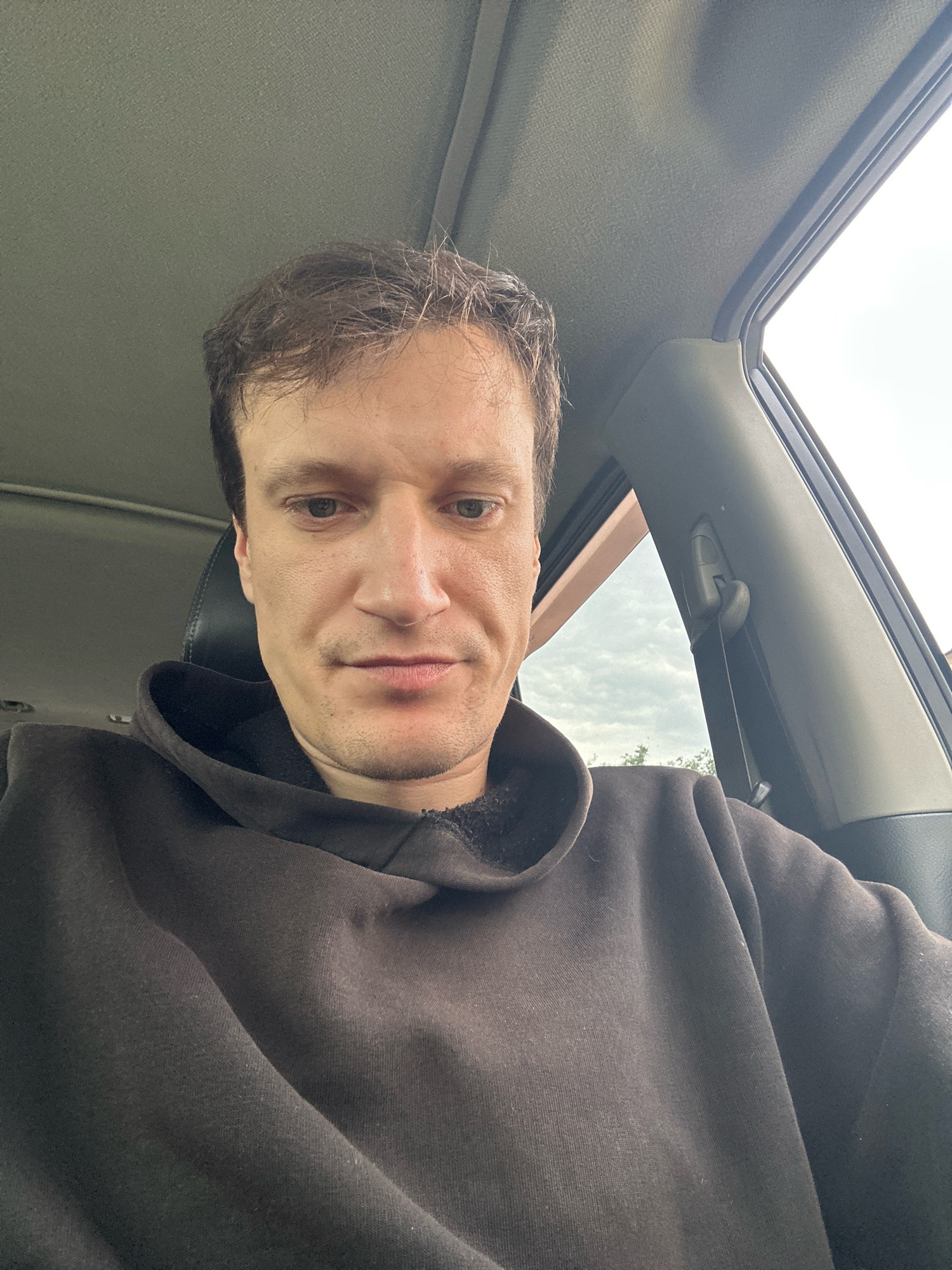 This is a selfie of a man taken inside a car. He is wearing a dark brown hooded sweatshirt. The man has short, brown hair and appears to be looking down. The background shows the interior of the car, including the ceiling, a window, and part of a seat. Outside the window, trees and a cloudy sky are visible. The location is identified as Makiv, Ukraine.