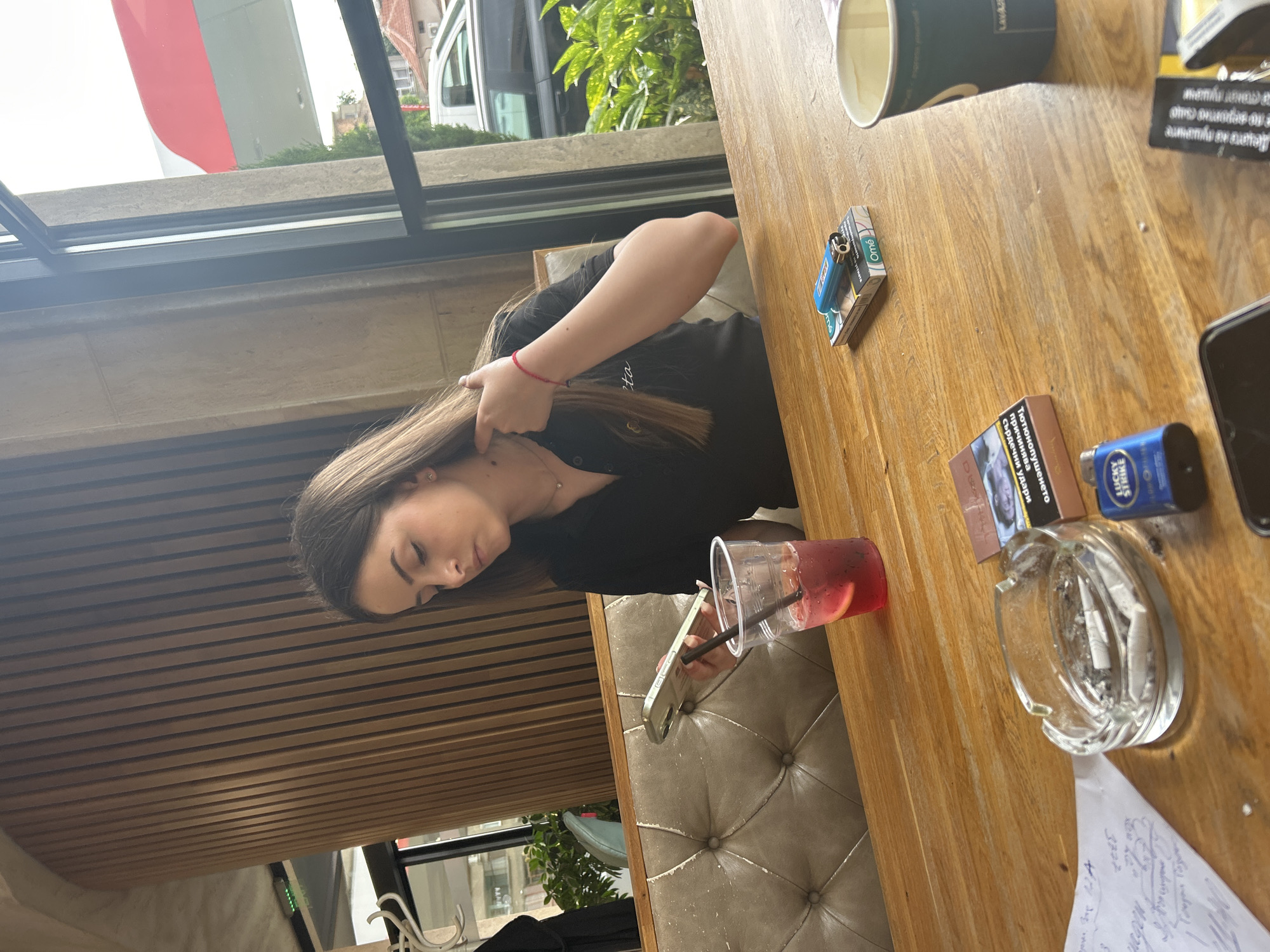 The image depicts a young woman sitting at a wooden table at an outdoor cafe or restaurant. She is holding her hair and looking down at her phone. The table contains a glass of red liquid with ice and a straw, an ashtray with cigarette butts, a pack of Lucky Strike cigarettes, a pack of Omé cigarettes, a lighter, a cup, a phone, and a piece of paper with handwriting on it. The table is situated near a window, through which one can see a street with vehicles and greenery. The handwriting on the piece of paper has the text "3372" in it. Some of the text on the Lucky Strike cigarette pack reads "Тютюнопушенето причинява сърдечни удар" which roughly translates to "Smoking causes heart attack". The overall impression is a casual setting where the woman is relaxing and occupied with her phone, surrounded by typical cafe items. It's unclear if the location is Kazanlak, Bulgaria without further evidence.