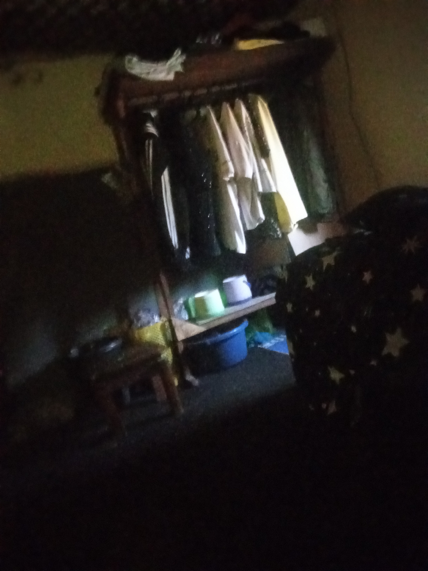 The image depicts a dimly lit interior scene, possibly a bedroom or storage area. In the center, a wooden shelf unit holds several items. On the top shelf, there are some white folded garments and what appears to be a yellow object. Below this, a clothing rack is filled with various garments, mostly light-colored, with some dark items also visible. To the left of the shelf unit, a small wooden stool with a woven seat is placed on the floor. On a lower shelf of the unit, there are two plastic containers, one light green and the other white, and a blue bucket is situated on the floor in front of the shelf. To the right, a large dark object covered in a white star pattern is partially visible. The overall lighting is poor, with shadows obscuring many details. The environment appears to be a domestic setting, possibly in Jos, Nigeria, given the provided context. There are no individuals clearly visible or identifiable in the image. The scene suggests a room that is not actively being used at the moment of capture, with items stored and arranged on shelving.