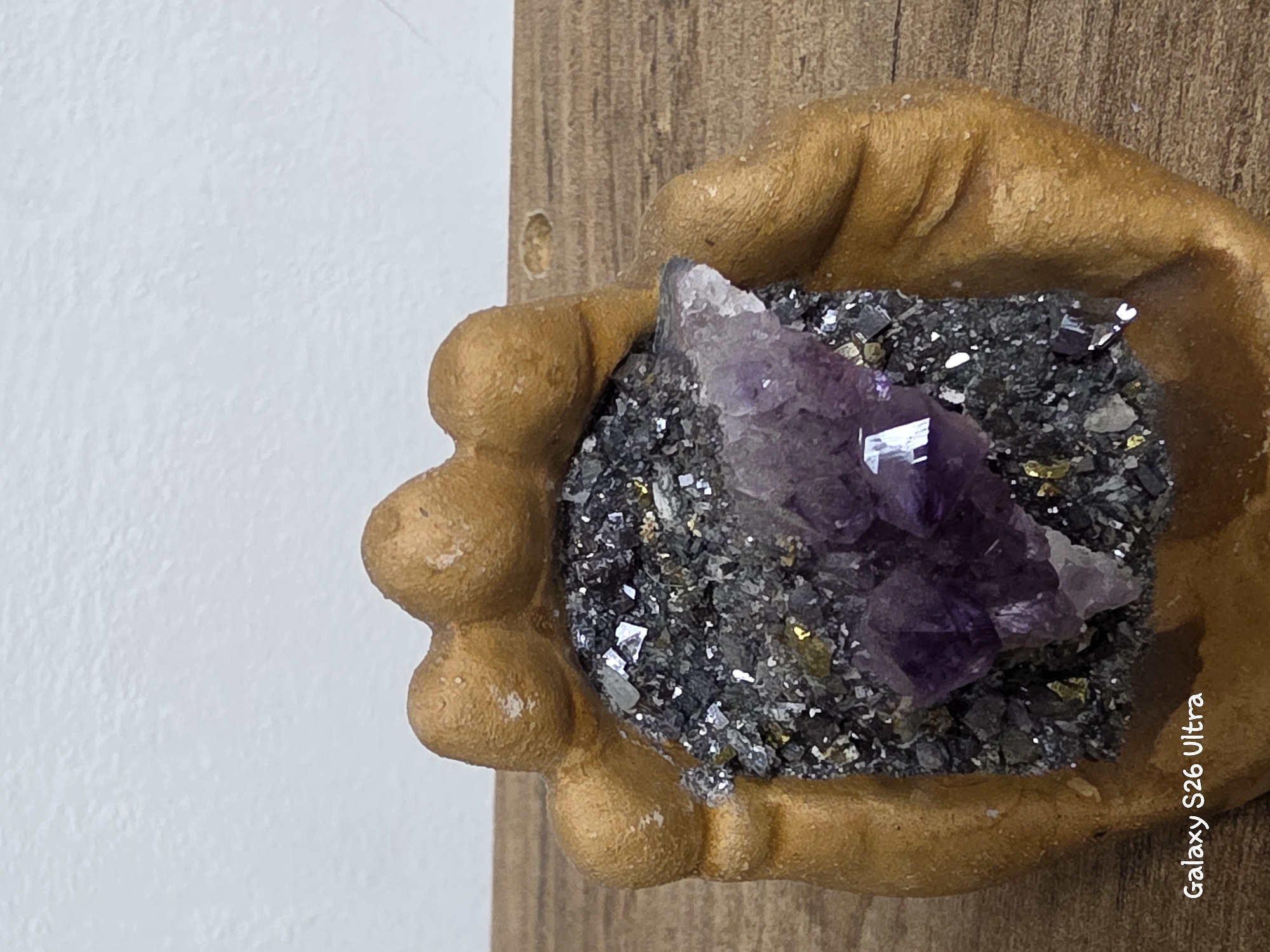 The image displays a close-up of a decorative object resembling a golden hand cradling a cluster of dark gray and purple crystals. The hand is stylized and appears to be made of a matte, textured material. The crystals are multifaceted, with the purple ones being more prominent and centrally located, and the dark gray ones forming a base. Some small, metallic-colored flecks are interspersed with the gray crystals. The object is placed on a wooden surface, possibly a table, with a plain, light-colored background behind it. The bottom left corner of the image contains the text "Galaxy S26 Ultra". The scene is indoors. No information regarding the city of Plovdiv, Bulgaria is present in the image.