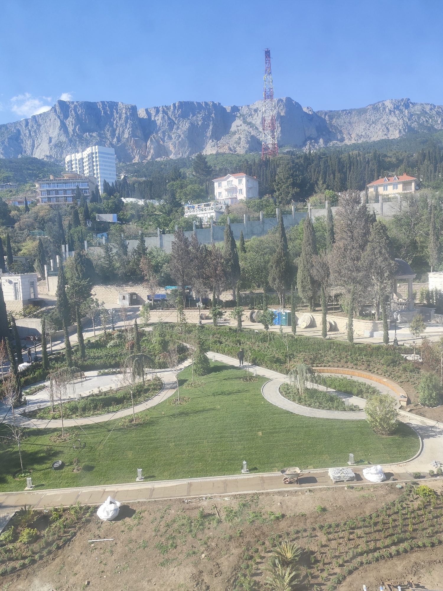 The image depicts a park situated in an unknown location, likely a city with mountainous terrain. The scene features a well-maintained park with manicured lawns and a network of paved pathways. A single individual is walking on one of the paths within the park. Adjacent to the park, there is an area with cultivated plants and construction materials, including a wheelbarrow, stone slabs, and white bags. In the background, there is a series of buildings and a red and white communication tower on the slopes of a mountain range. The sky is clear and blue.