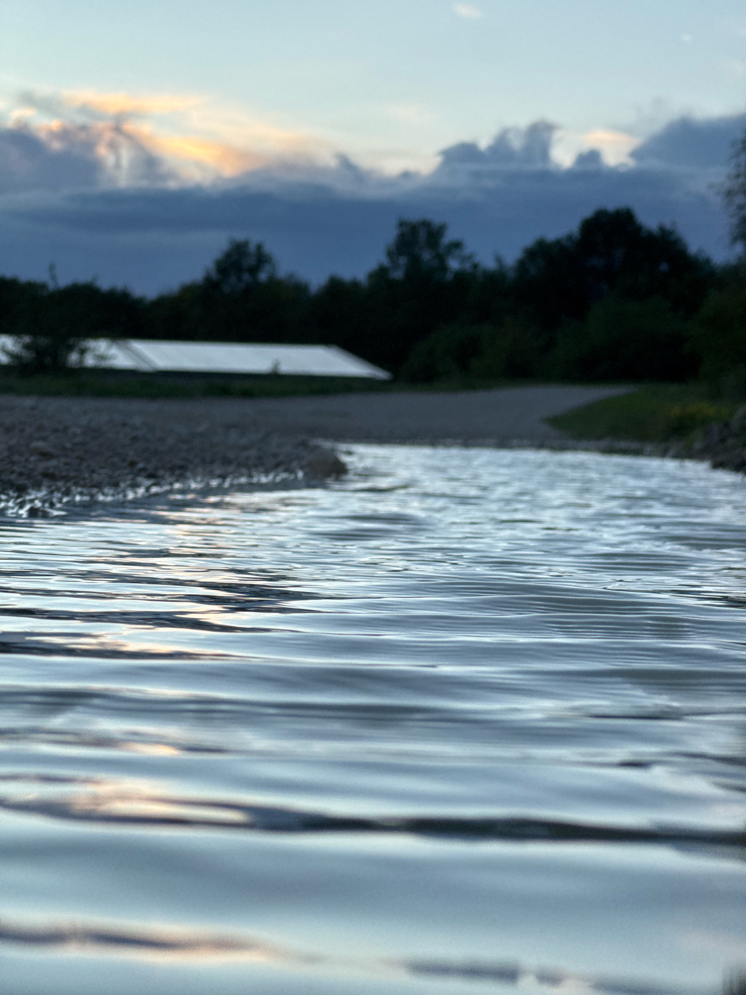The image depicts a shallow body of water in the foreground, with gentle ripples reflecting the sky. Beyond the water, a pebbled shoreline is visible, leading to a dark line of trees. In the background, partially obscured by trees, is a large, white, angled structure that appears to be a solar panel array. The sky above is a pale blue, with dark, dramatic clouds that are illuminated by soft, golden light, suggesting either sunrise or sunset. The scene is serene and natural, with a man-made element (the solar panels) integrated into the landscape. The location is identified as Wettstetten, Germany. There are no people or animals visible in the image.