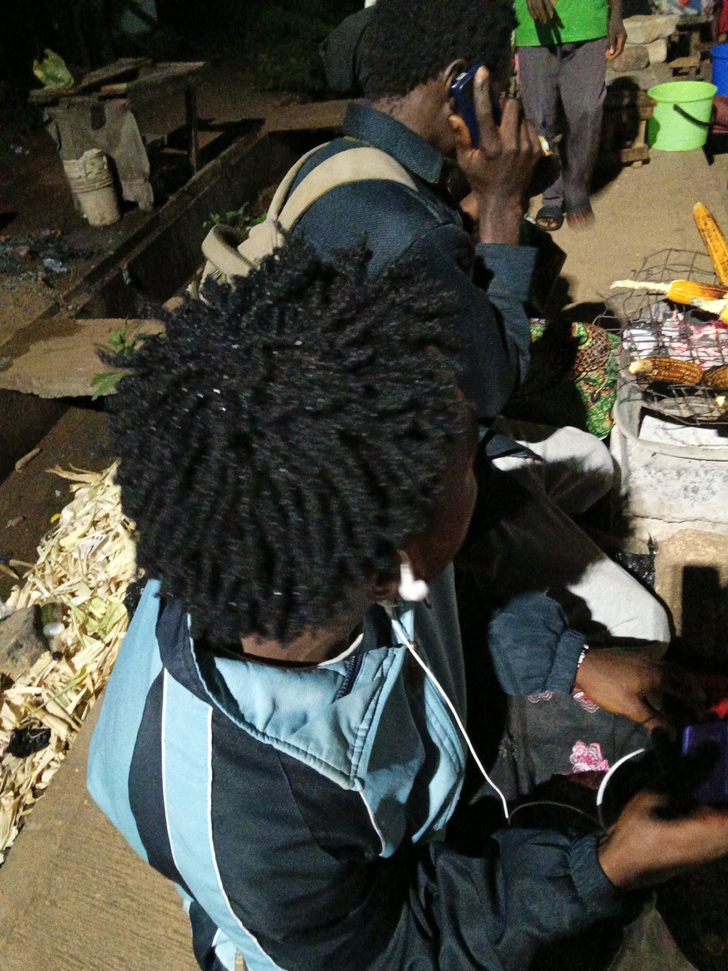 The image depicts a street scene in Bukuru, Nigeria, at night. In the foreground, a young person with dark, braided hair wears a blue and black jacket with light blue and white stripes. They are holding a purple object in their right hand and a white cord is visible. Their head is turned to the right, and their face is partially obscured. To the left, a pile of dried corn husks and stalks is on the ground. In the background, a person in a dark blue jacket talks on a blue cell phone. Another individual in a green shirt and grey pants stands behind them. A metal grill with corn on it is visible, along with a green bucket and wooden structures. The ground is dirt and pavement.