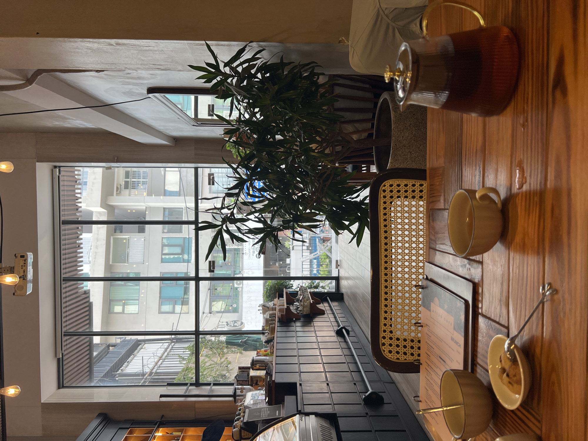 The photograph depicts the interior of a café, likely in Bắc Nha Trang, Vietnam, as suggested by the text “342 6748” on a street-facing sign visible through the expansive window. The café’s aesthetic includes large, black-framed windows that offer a view of the city. Through the window, one can discern apartment buildings and a street scene.
The foreground features a wooden table, upon which sits a clear teapot filled with tea, two teacups, and a small dish with cookies or pastries and a spoon. A menu card is also on the table.
A cane-backed chair is visible in the midground. A potted plant stands behind the chair, partially obscuring the view outside. A counter is positioned along the base of the window. The atmosphere is calm and subdued, with a focus on enjoying tea in a modern, urban setting.
