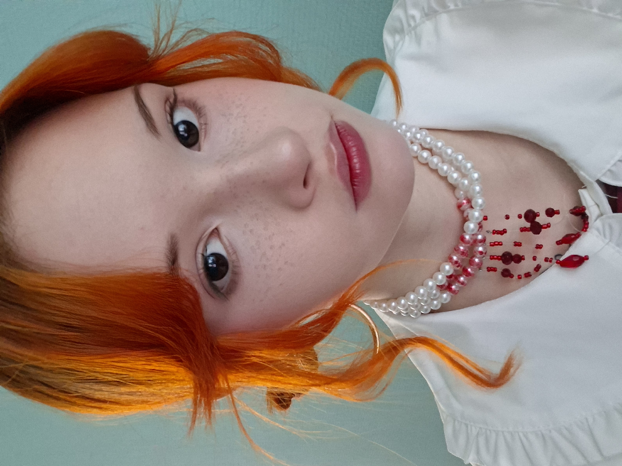 A close-up, diagonally oriented portrait features a young woman with vibrant, bright orange-red hair. Her fair skin is notable for numerous freckles scattered across her nose and cheeks. She has wide, dark eyes that gaze directly at the viewer, and her lips are painted with a glossy, reddish-pink color. She is wearing a white blouse, possibly with ruffled detailing on the collar or neckline. Adorning her neck are two distinct necklaces: a double strand of white or cream-colored pearls, and a separate, more intricate red beaded necklace featuring various sizes and shades of red beads. The background is a plain, light blue-green surface, suggesting an indoor setting. The overall impression is a direct and artful portrait. No text is visible in the image. This media file originates from Chaikovsky, Russia.