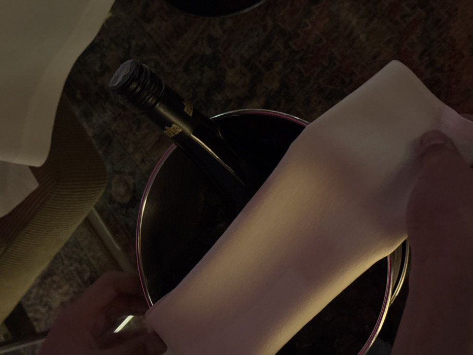 The image depicts an indoor scene within a dining establishment in Yerevan, Armenia. The central subject is a dark-colored wine bottle positioned upright within a metallic ice bucket, which appears to contain ice and liquid. A white, folded cloth or napkin is draped over the rim of the ice bucket and partially covers the bottle. Two human hands, likely adult, are visible in the lower foreground, holding the edges of this white cloth, suggesting an act of handling or service. The wine bottle displays a gold-toned label with faintly visible, largely unidentifiable text, and its neck is secured with a dark, ribbed closure. The background includes a fragment of a light-colored upholstered chair, a section of a white tablecloth, and a dark, patterned carpet or rug on the floor. The environment is illuminated by dim, artificial lighting. No other individuals or specific identifying architectural features of the exact venue are discernible.