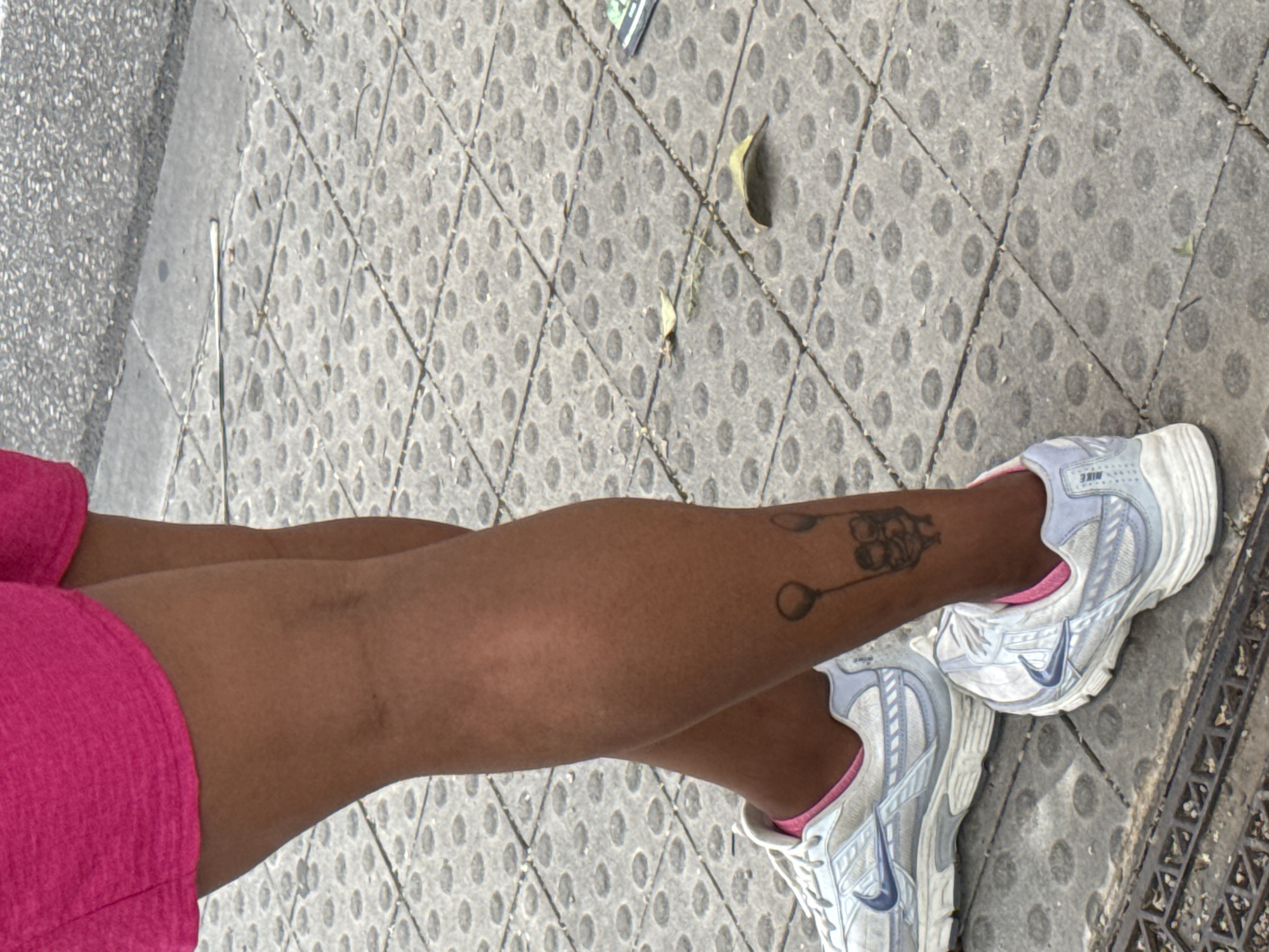 A medium shot captures a person's lower legs and feet standing on a textured, grey tiled sidewalk. The person is wearing a bright pink skirt or shorts and light blue and white Nike sneakers with pink socks. A small, dark tattoo of two figures holding balloons is visible on the lower left leg. The sidewalk tiles are arranged in a grid pattern, with raised circular bumps on each tile. There are a few scattered dry leaves on the ground. The scene appears to be outdoors in an urban setting, likely in Gandia, Spain, given the context of the inquiry.