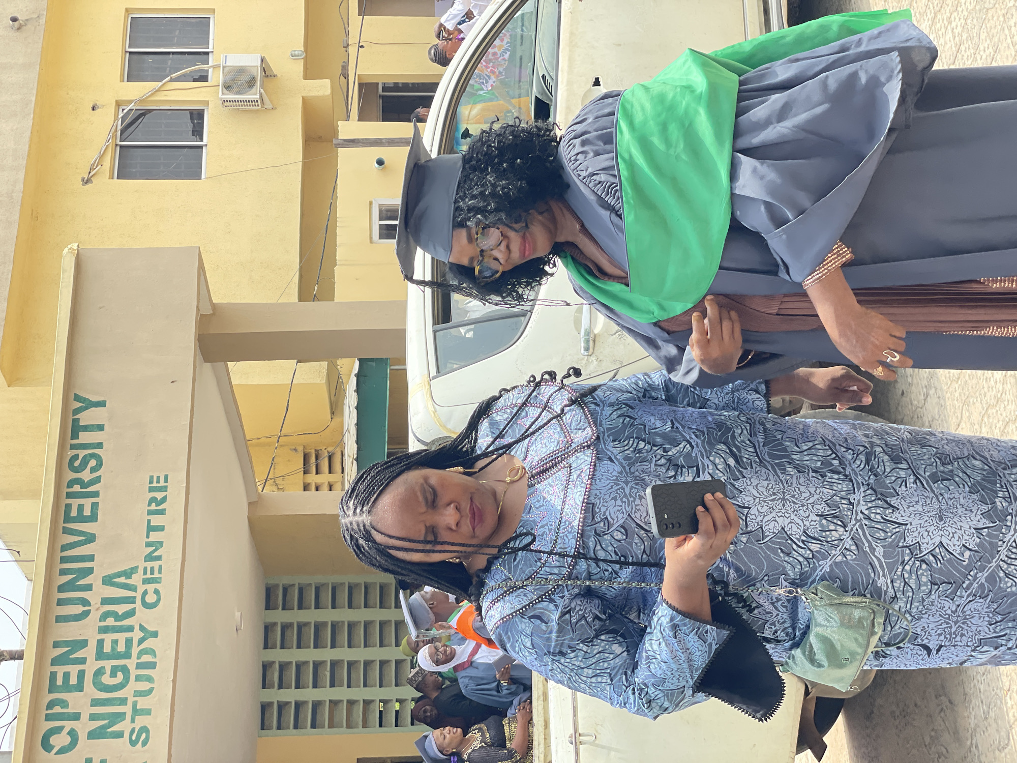 The photo captures two women in what appears to be a celebratory setting. A woman wearing a graduation gown (grey with a green sash) and mortarboard cap stands near another woman who holds a cell phone. Both women are dark skinned. The woman holding the cell phone is dressed in a blue patterned dress.
The background features a beige building with the words "Open University Nigeria Study Centre" written in green. An air conditioning unit is visible on the building, along with windows with barred grilles.
Additional people are visible in the background.
Based on the sign on the building, the location is likely at the Open University Nigeria Study Centre. As the sign does not state the specific location of the university, the claim that it is in Lokoja cannot be confirmed.
