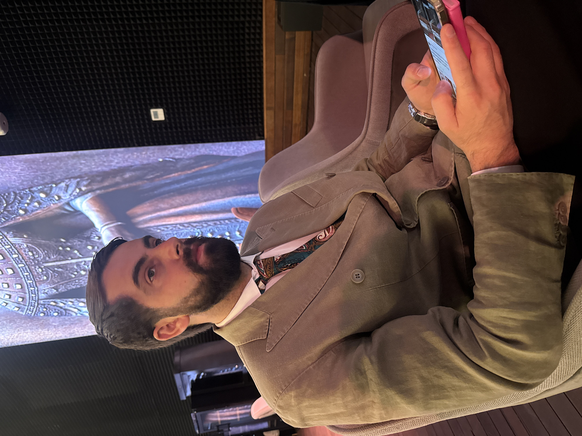 A man with a beard is sitting in a mauve-colored chair, looking to the left while holding and viewing his smartphone. He is wearing a tan-colored suit jacket with a patterned tie. A large screen displaying what appears to be an image of a film or television scene is visible behind him. The background also features soundproofing panels. Given the mention of Yerevan, Armenia, this scene likely takes place inside a cinema or a media production studio within the city.