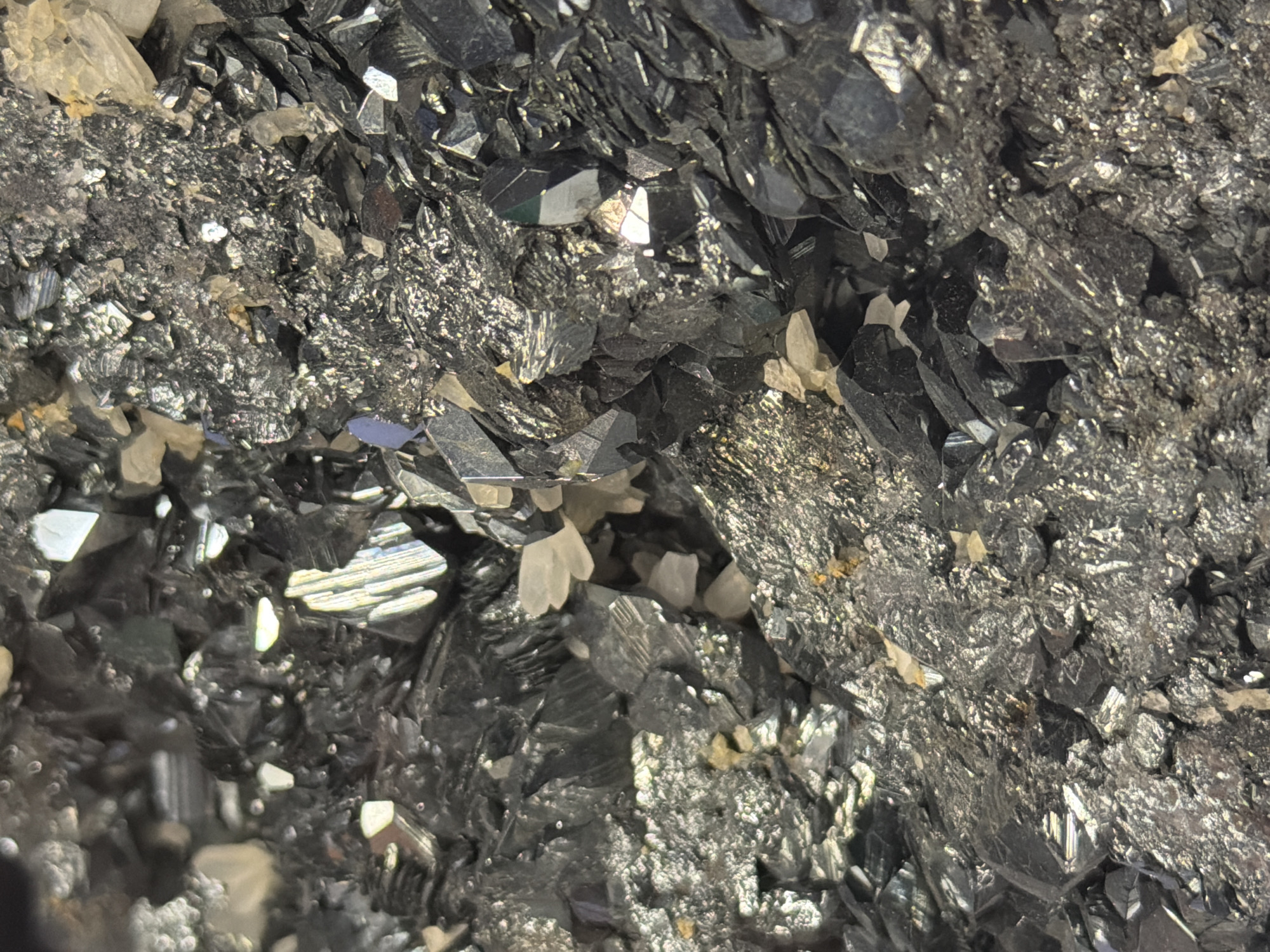 The image shows a close-up, macro view of a dark, crystalline mineral formation. The crystals are primarily black and metallic, with some appearing to be hexagonal or prismatic in shape. There are also lighter-colored, translucent crystals interspersed among the darker ones. The texture is rough and irregular, with many facets reflecting light. The scene is indoors, possibly in a museum or collection, given the nature of the subject. No specific location in Fort Lauderdale, United States of America can be identified from the image.