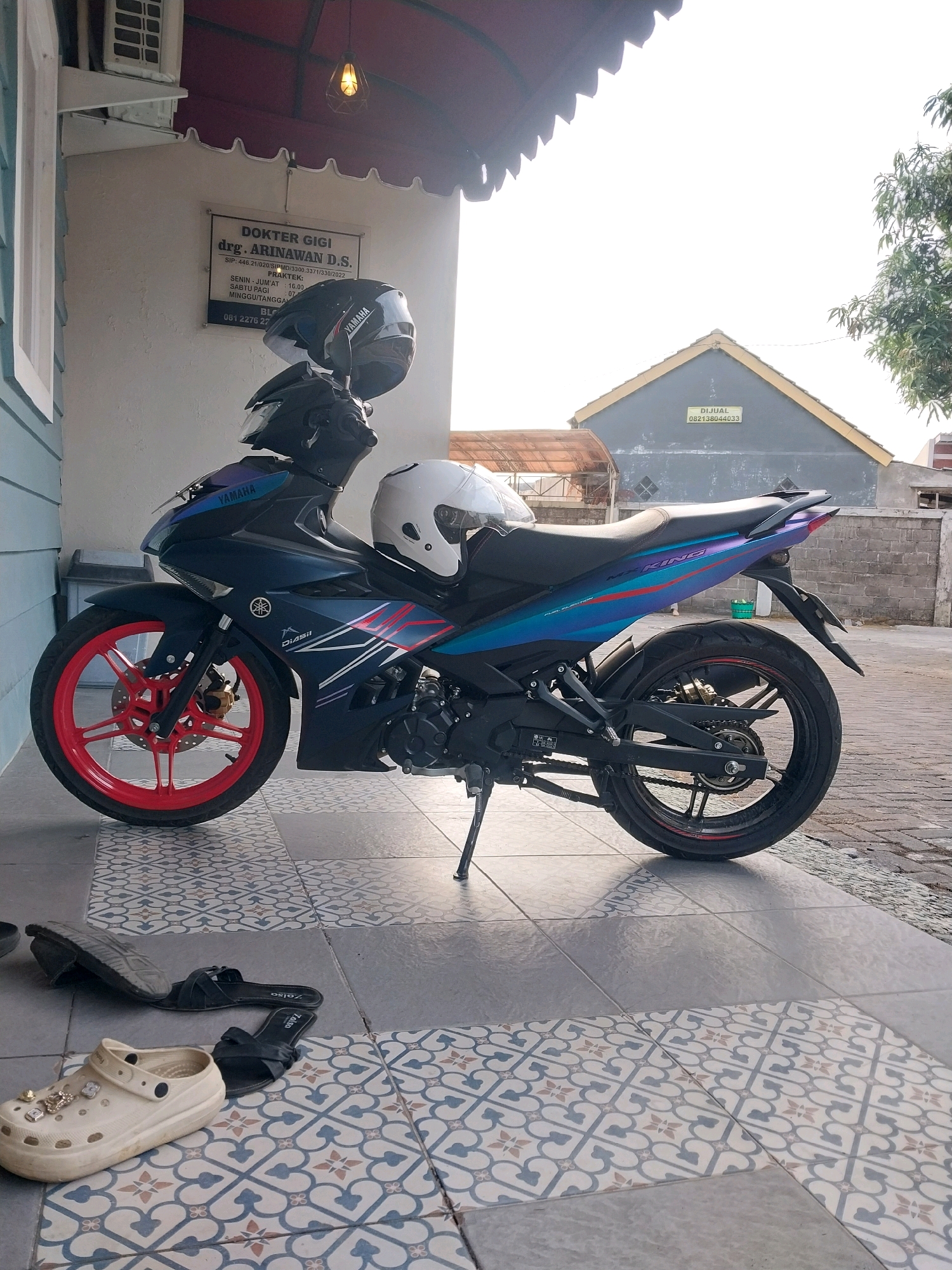 A dark blue Yamaha motorcycle is parked on a tiled patio in front of a building in Magelang, Indonesia. The motorcycle has red rims and two helmets are placed on it, one black and one white. To the left of the motorcycle, there is a light blue wall with a white air conditioner unit mounted on it. A sign on the wall reads "DOKTER GIGI DRG. ARINAWAN D.S." and provides practice hours. On the patio, there are two pairs of black sandals and a pair of beige Crocs adorned with charms. In the background, a gray building with a yellow roof has a sign that reads "DIJUAL 082138044033". The sky is bright and overcast.