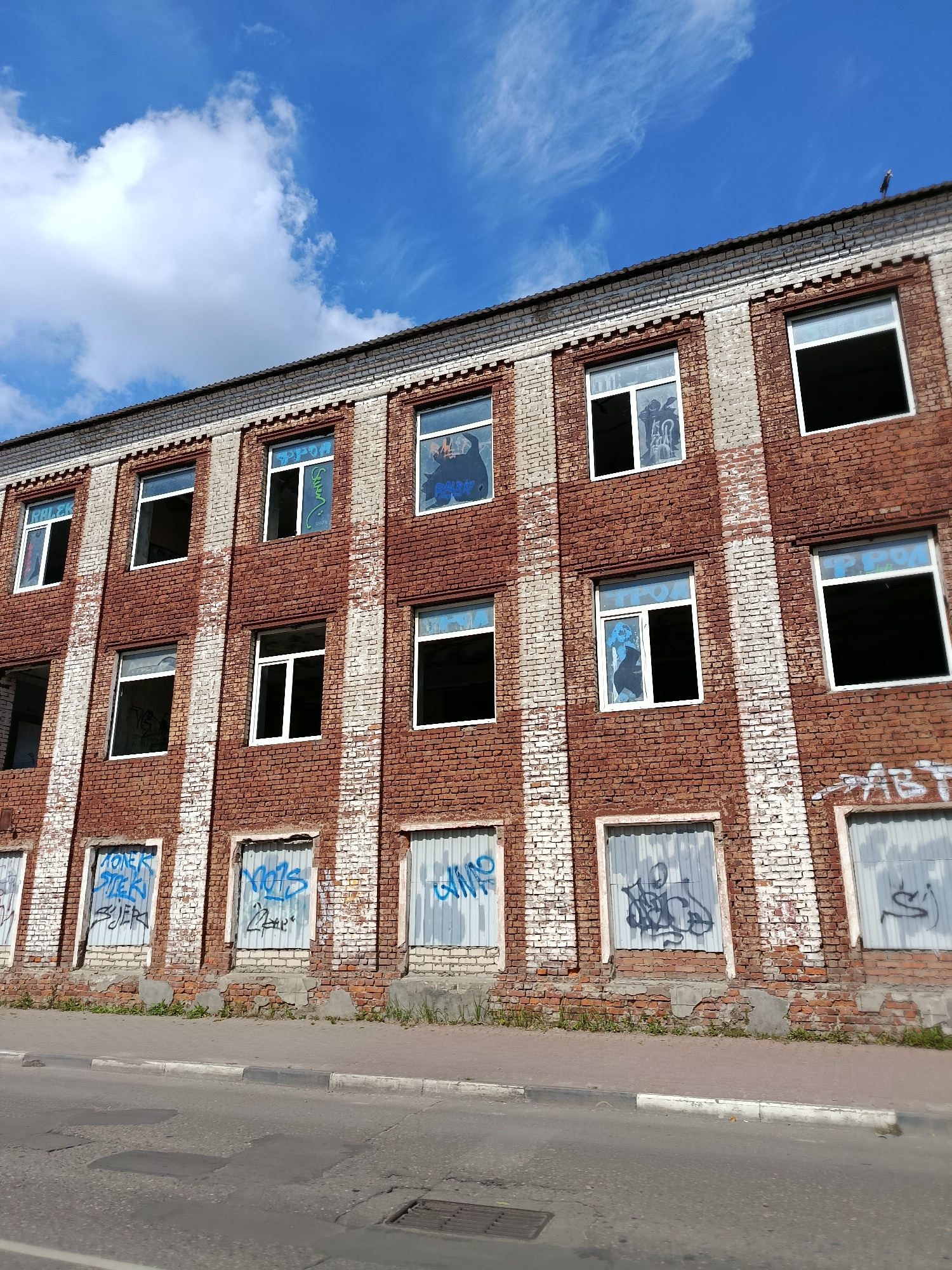 An abandoned brick building with graffiti is visible in Orekhovo-Zuyevo, Russia, under a blue sky with scattered clouds. The building has three floors, each with multiple windows. Some windows are broken or boarded up with corrugated metal sheets, which are also covered in graffiti. The brickwork is weathered, with some areas painted white that are peeling away. The ground in front of the building is a paved sidewalk next to a street with visible asphalt repairs. In the distance, a single bird is perched on the roofline of the building.
