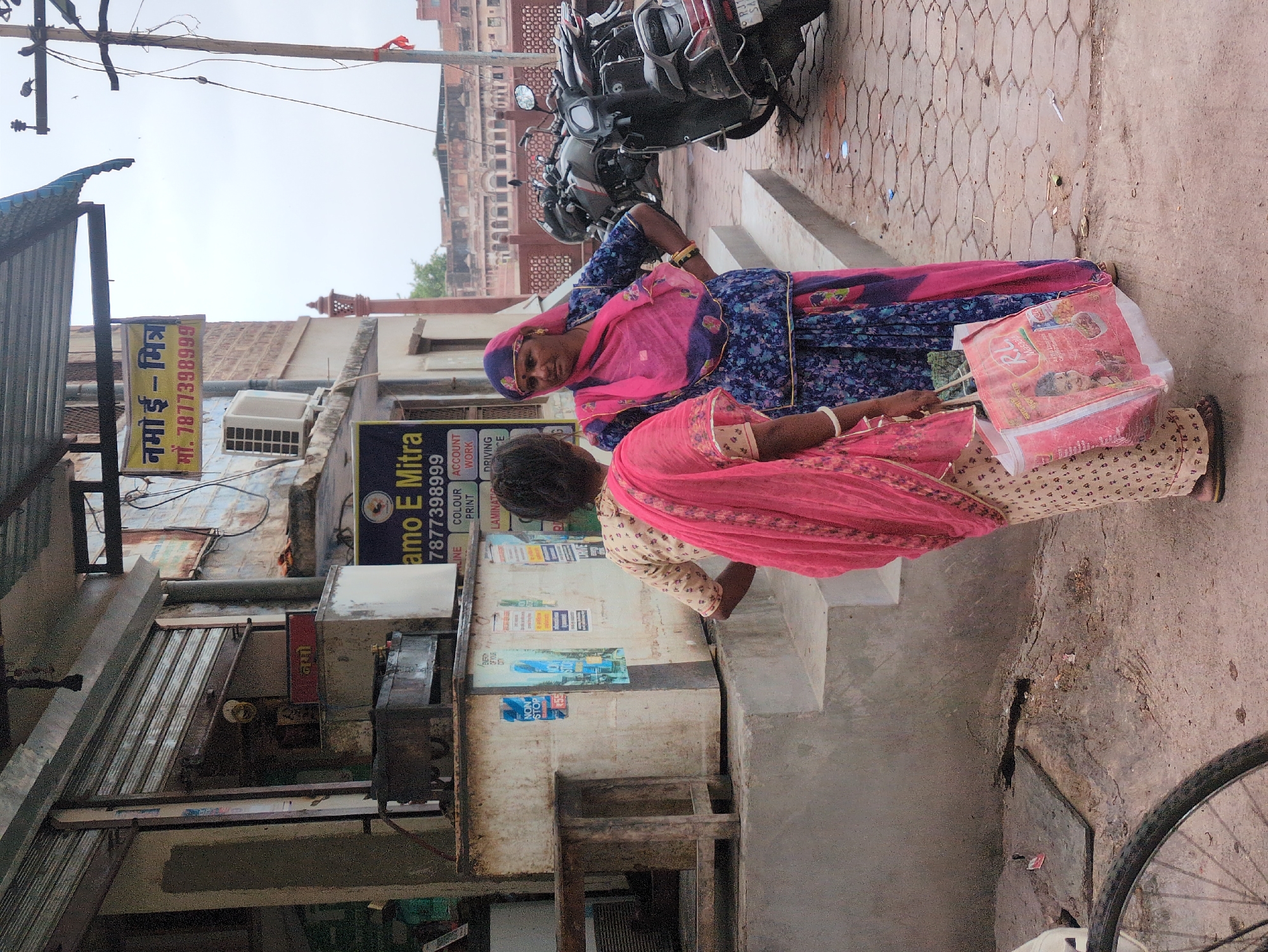 This is a medium-shot, eye-level, outdoor photograph taken in Jodhpur, India. The scene depicts two women interacting on a paved walkway in front of a building. The woman on the right, wearing a blue floral-print sari and a pink headscarf, is facing left and speaking to the woman on the left. The woman on the left, her back mostly to the camera, wears a cream-colored sari with a pink border and a pink scarf draped over her shoulders. She is holding a pink and white reusable shopping bag featuring an image of a young girl and the word "Masala." Behind the women, a row of motorcycles is parked along the walkway. The building behind them appears to be a shop with a metal roll-up door. A sign above the shop reads "namo e-mitra" in Hindi and English, with a phone number "9877398999." Another sign below that advertises services such as "account," "work," and "driving." The ground is paved with hexagonal tiles. A portion of a bicycle wheel is visible in the lower-left corner of the frame. The sky is overcast.