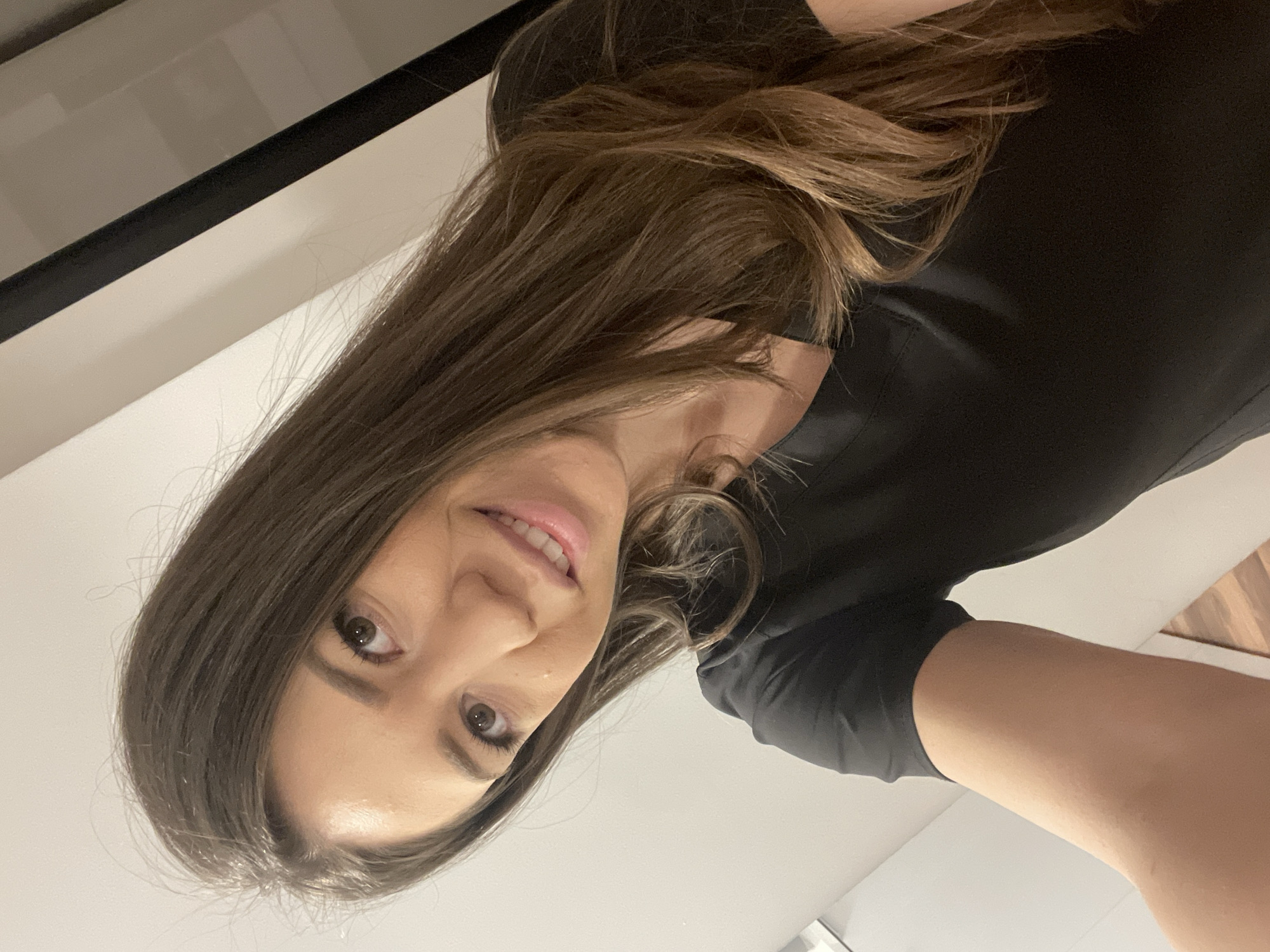 A woman is posing for a selfie. She has brown hair, fair skin, and dark eyes. She is wearing a black shirt. The background appears to be an interior setting with white walls and a dark-framed window or door visible in the upper left corner. It is not possible to determine the location in Sofia, Bulgaria from this image alone.