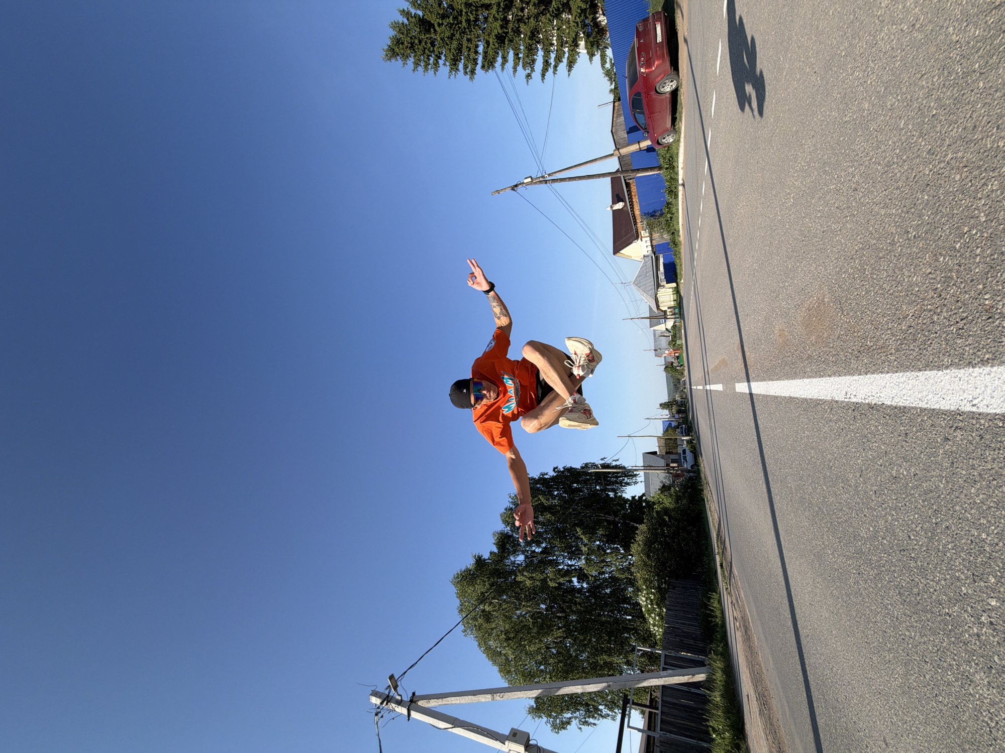 A man wearing an orange T-shirt, black shorts, and a black baseball cap is jumping in the air. He is on a street in Chaikovsky, Russia, with a clear blue sky above. A red car is parked alongside the road with a blue fence. Utility poles, trees, and buildings line the road. The road is paved with a white dividing line. The man's arms are extended.