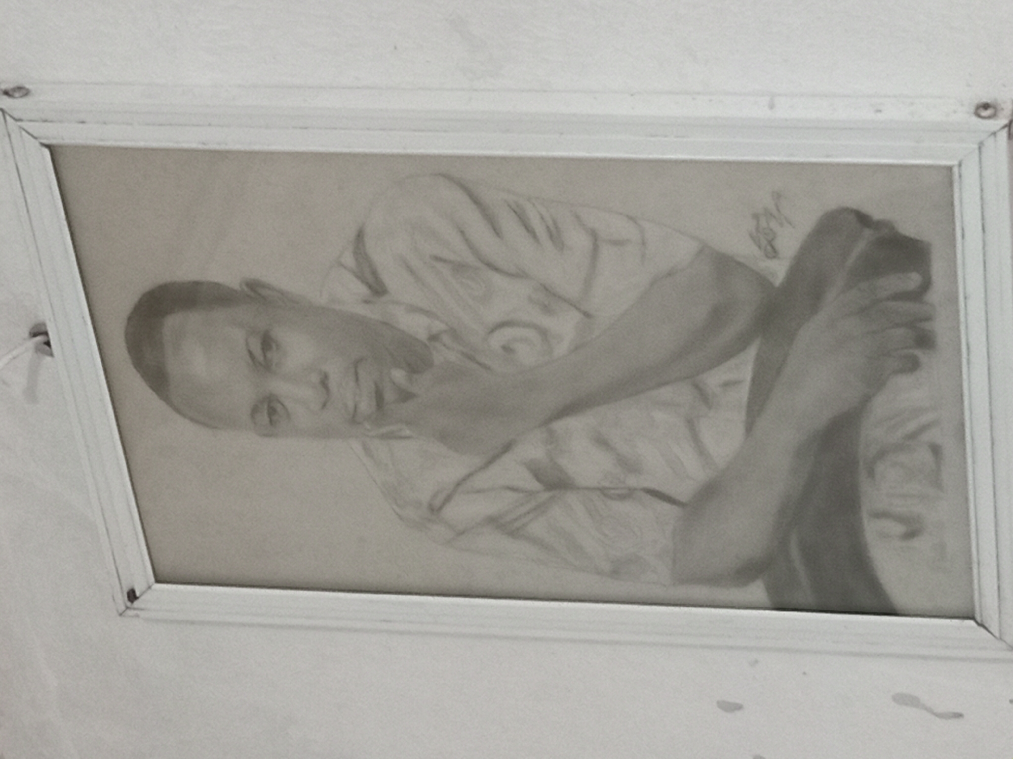 The media file presents a framed monochromatic drawing, likely a pencil or charcoal sketch, displayed on a plain, light-colored wall. The drawing features a single subject, an adult male, depicted in a relaxed, reclining pose. His head is turned towards the viewer, with a calm, possibly contemplative expression. One hand rests near his chin, while his other arm is visible, and his legs are bent, suggesting a seated or propped-up position. The artwork showcases detailed shading that defines facial features, clothing folds, and body contours.
In the upper right quadrant of the drawing, a dark, stylized signature or initials, which appears to be a cursive 'K' or similar, is visible. The drawing is enclosed within a simple, light-colored, possibly white, wooden frame. The frame shows minor signs of wear, including small holes or hooks at its top corners, indicating how it is mounted. The wall itself is a uniform off-white or light grey, exhibiting some subtle texture or small, irregular marks. The photograph of the artwork is slightly rotated, showing the framed piece at an angle. The overall scene suggests an intimate indoor setting, potentially in Shagamu, Nigeria, showcasing a personal piece of art.