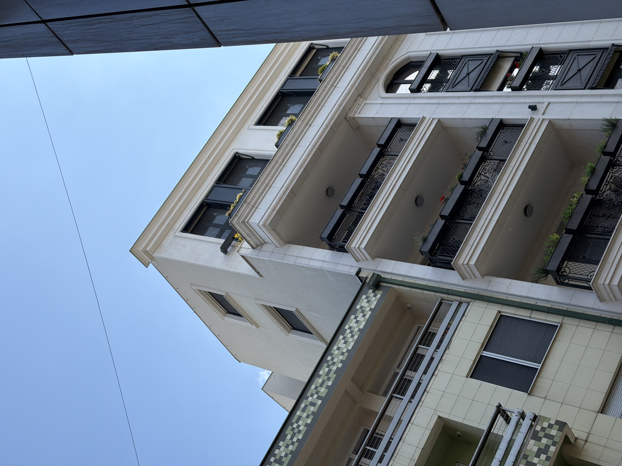 The image presents a low-angle view of multiple multi-story residential or mixed-use buildings under a clear blue sky. In the foreground, a prominent light cream-colored building features ornate classical architectural elements, including a heavy cornice at the roofline, textured facade, and multiple balconies with decorative black wrought-iron railings. Each balcony ledge holds black planter boxes containing green foliage and some red flowers. Windows are dark-framed, some equipped with exterior screens. A tall, narrow section on the far right of this building displays an arched window at the top, followed by black decorative panels with visible red flowers in pots below. To the left, a slightly recessed building exhibits a lighter beige facade with a distinctive horizontal band of green and light green checkered tiles below its roofline. This building includes at least one balcony with simple grey metal railings and dark-framed windows, some with screens. A dark green downspout is visible where this building meets the more ornate one. Further back and to the upper left, another light-colored building with dark-framed windows is partially visible. A dark grey, paneled vertical structure partially frames the right side of the image. A thin, dark utility wire traverses the upper portion of the clear blue sky. No individuals are present in the scene. The depicted location is within the Vracar Urban Municipality, Serbia.