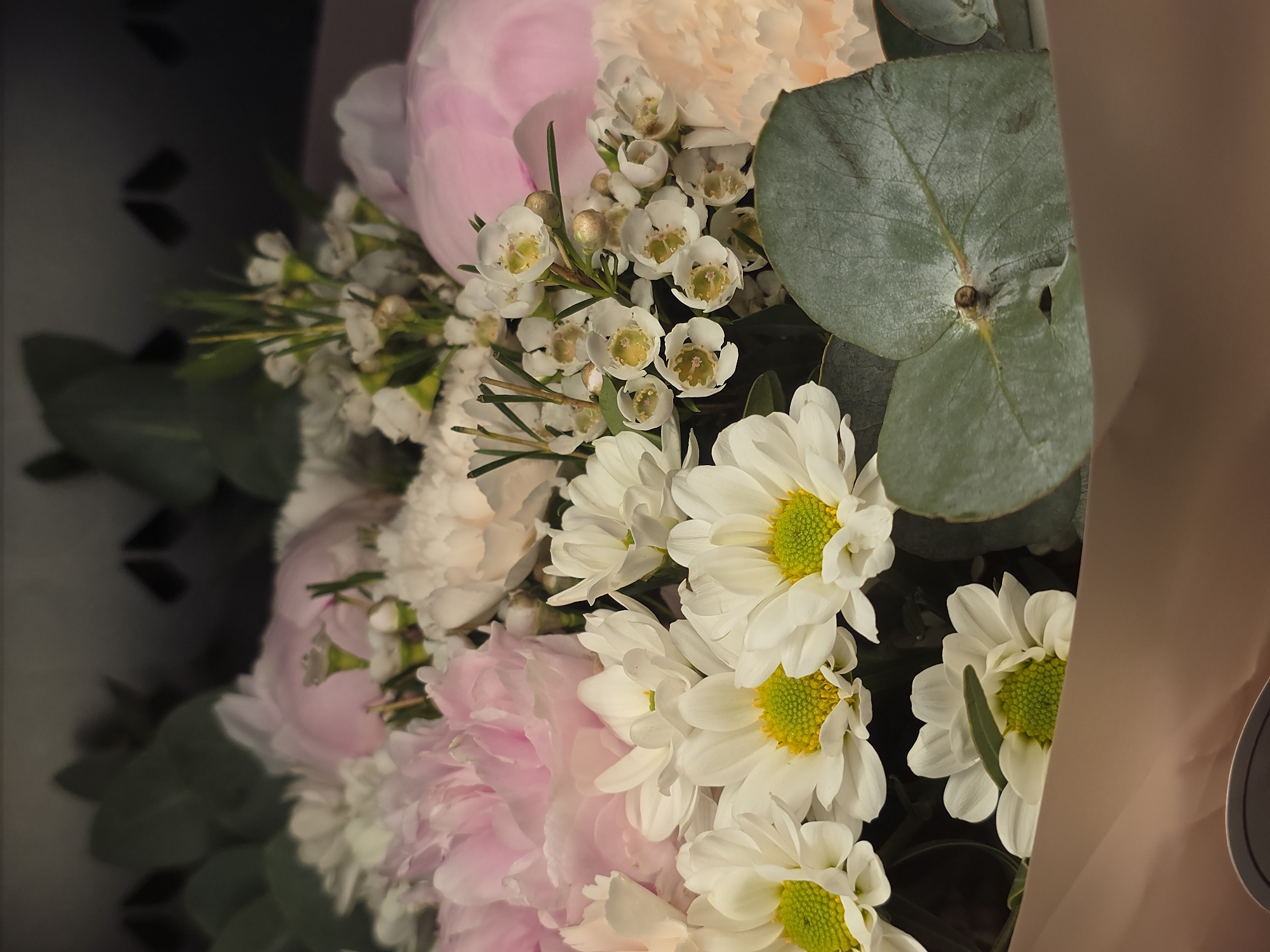 The image depicts a close-up of a bouquet of flowers. The arrangement includes pink peonies, white daisies with green centers, small white flowers resembling waxflowers, and eucalyptus leaves. The bouquet is partially wrapped in light brown paper. The location is unknown, and there is no indication of Warsaw, Poland. There are no people visible.