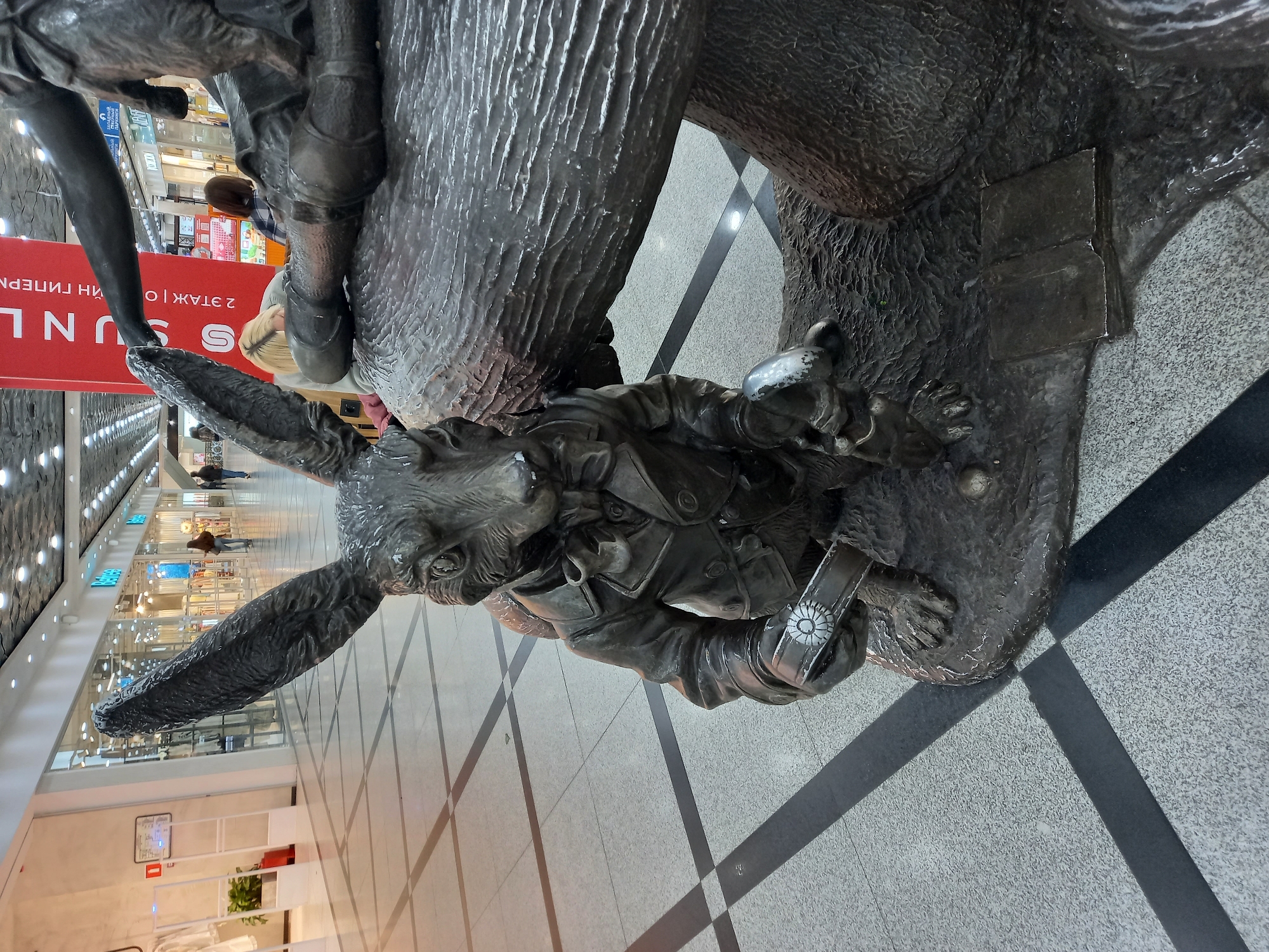 The image depicts a bronze sculpture of a rabbit character dressed in a jacket, holding a pocket watch. The sculpture is positioned on a textured base that appears to be part of a larger artistic installation, possibly representing a mushroom or a tree stump. A small, open book is visible on the base near the rabbit's feet. In the background, there are indications of a commercial setting, with shop windows and shoppers visible. A red banner with Russian text, including "UNL" and "2 ЭТАЖ" (2nd Floor), suggests this is a retail environment. The flooring is tiled with a geometric pattern of light and dark squares. The sculpture is located in the shopping center "UNIVERMAG BOLSHOY UNIVERSALNY MAGACIN" (Big Universal Store) in Yekaterinburg, Russia, which is known for its collection of Alice in Wonderland themed sculptures.